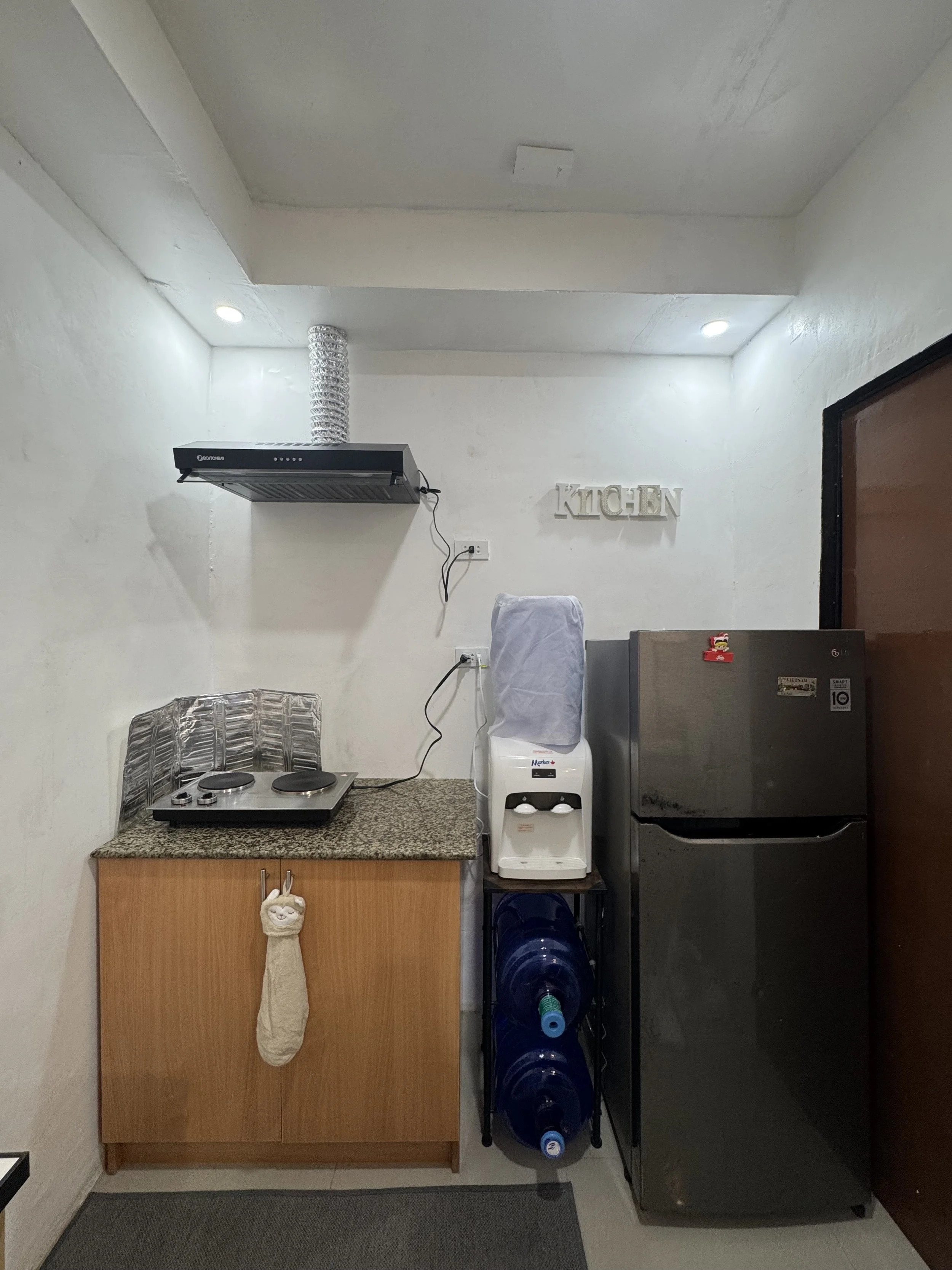 A compact kitchen corner with a wooden cabinet, a countertop with a two-burner stove, a water dispenser, a metallic water filter, and large blue water bottles underneath. The wall has decorative letters spelling 'KITCHEN' and the ceiling has recessed