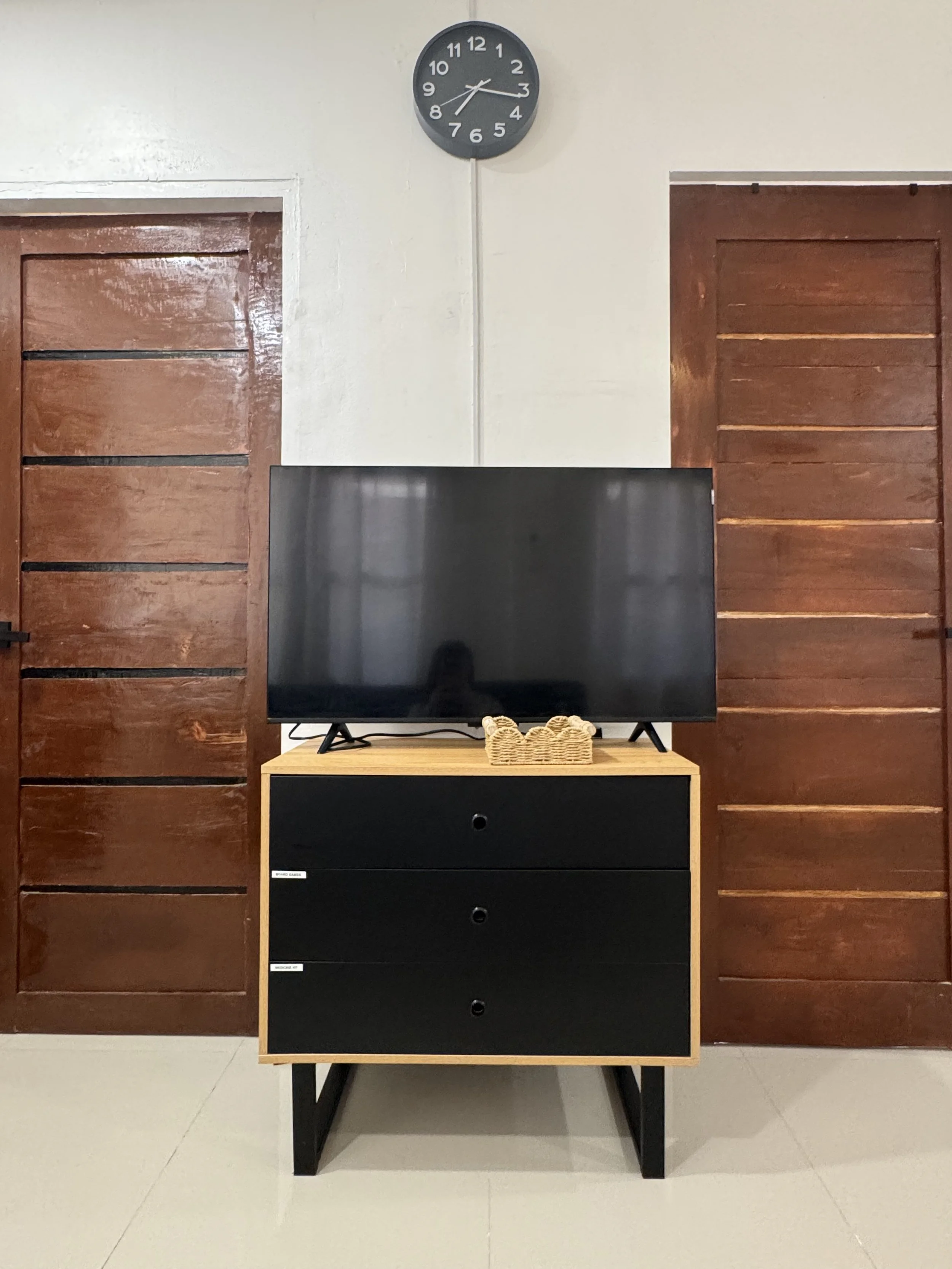 A modern black and wood TV stand with three drawers, a woven basket, and a flat-screen TV. Flanked by two wooden doors and a wall clock showing 2:15.