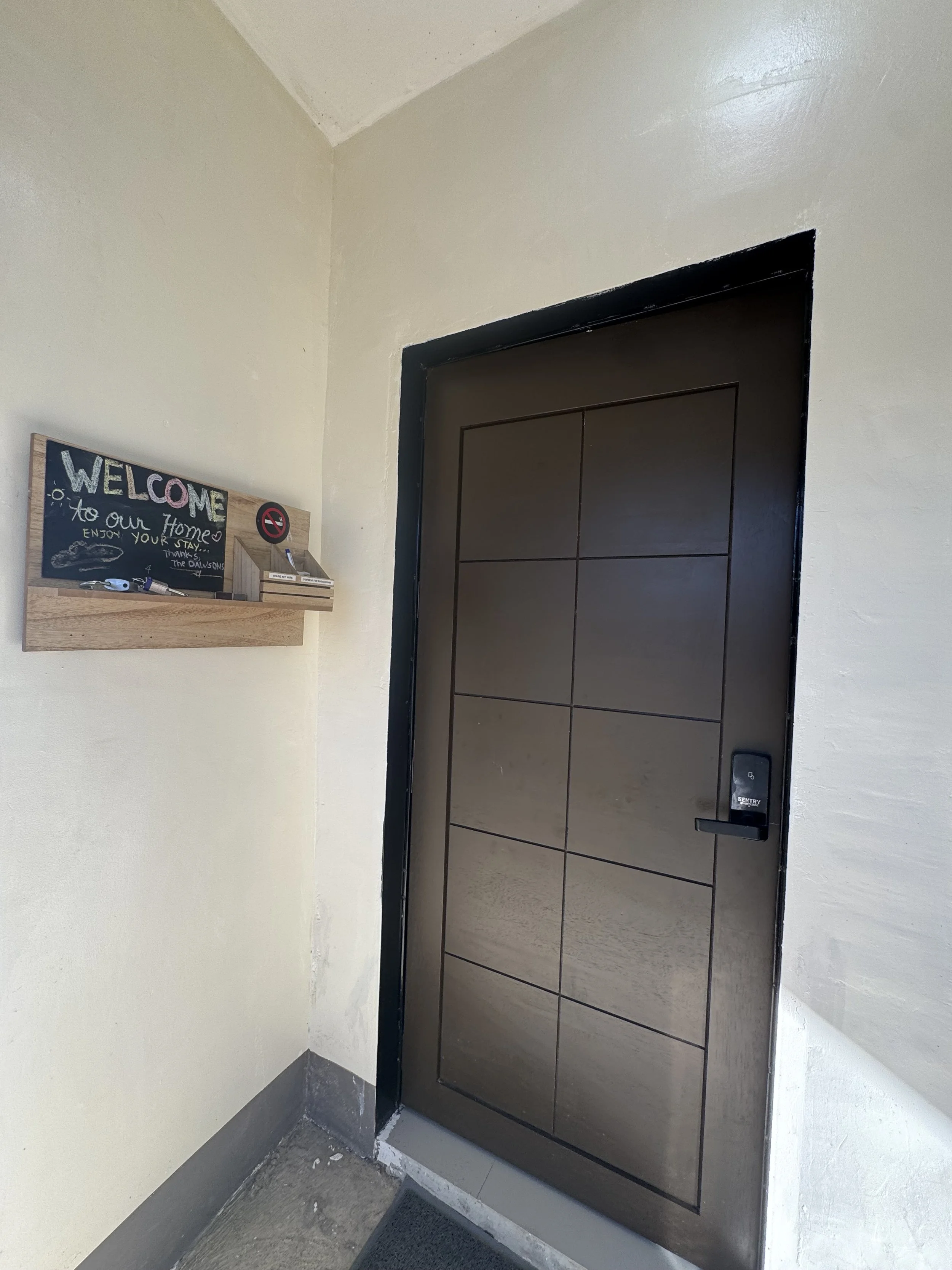 An elevator door with a black frame and brown panels. A keypad or intercom is mounted next to the door. A wooden chalkboard sign with colorful writing and a small box with a cigarette icon are on the wall next to the door.