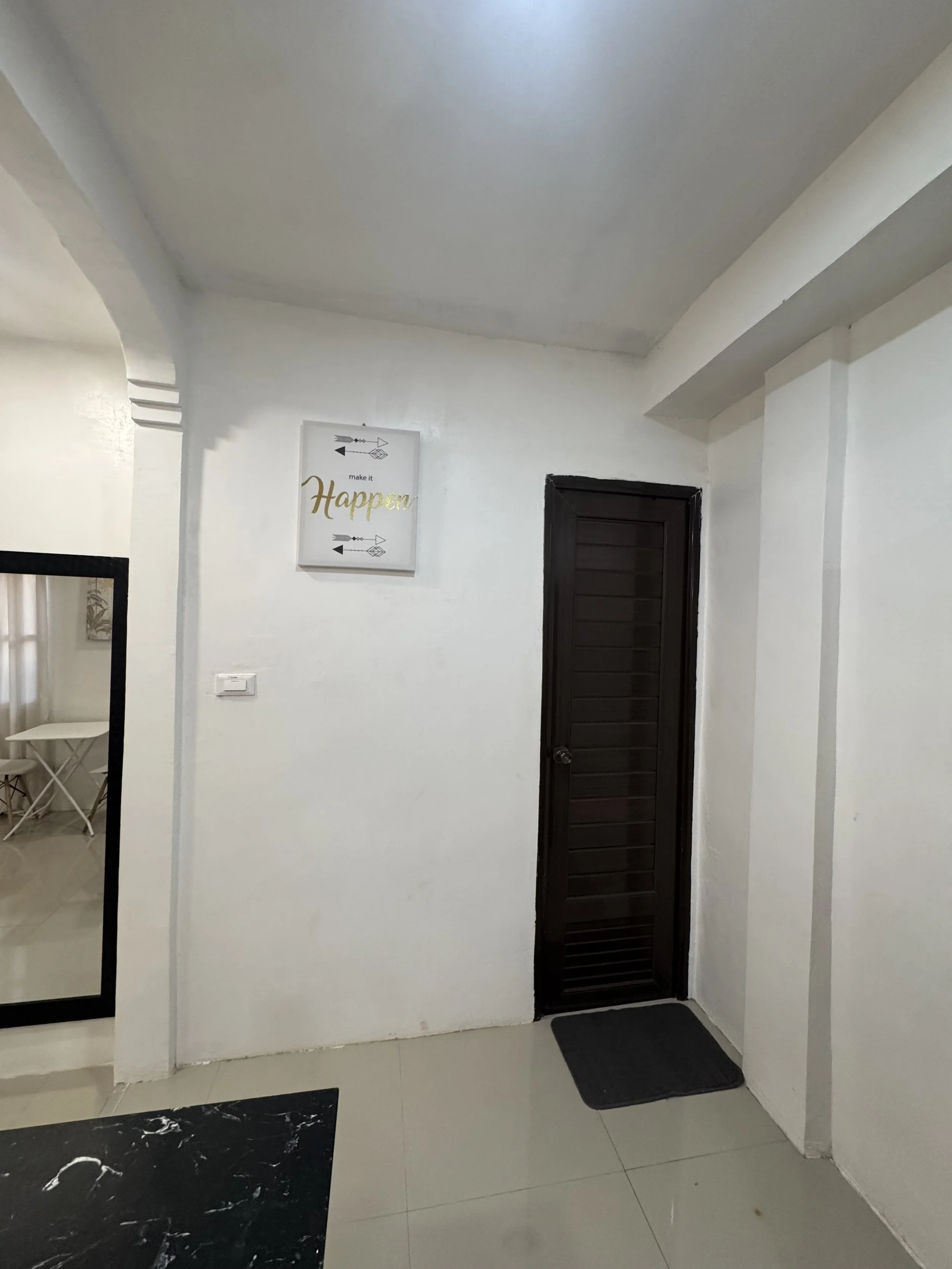 Indoor hallway corner with a black door, a wall-mounted motivational sign, a black doormat, and a part of a large mirror reflecting a chair and side table in a room.