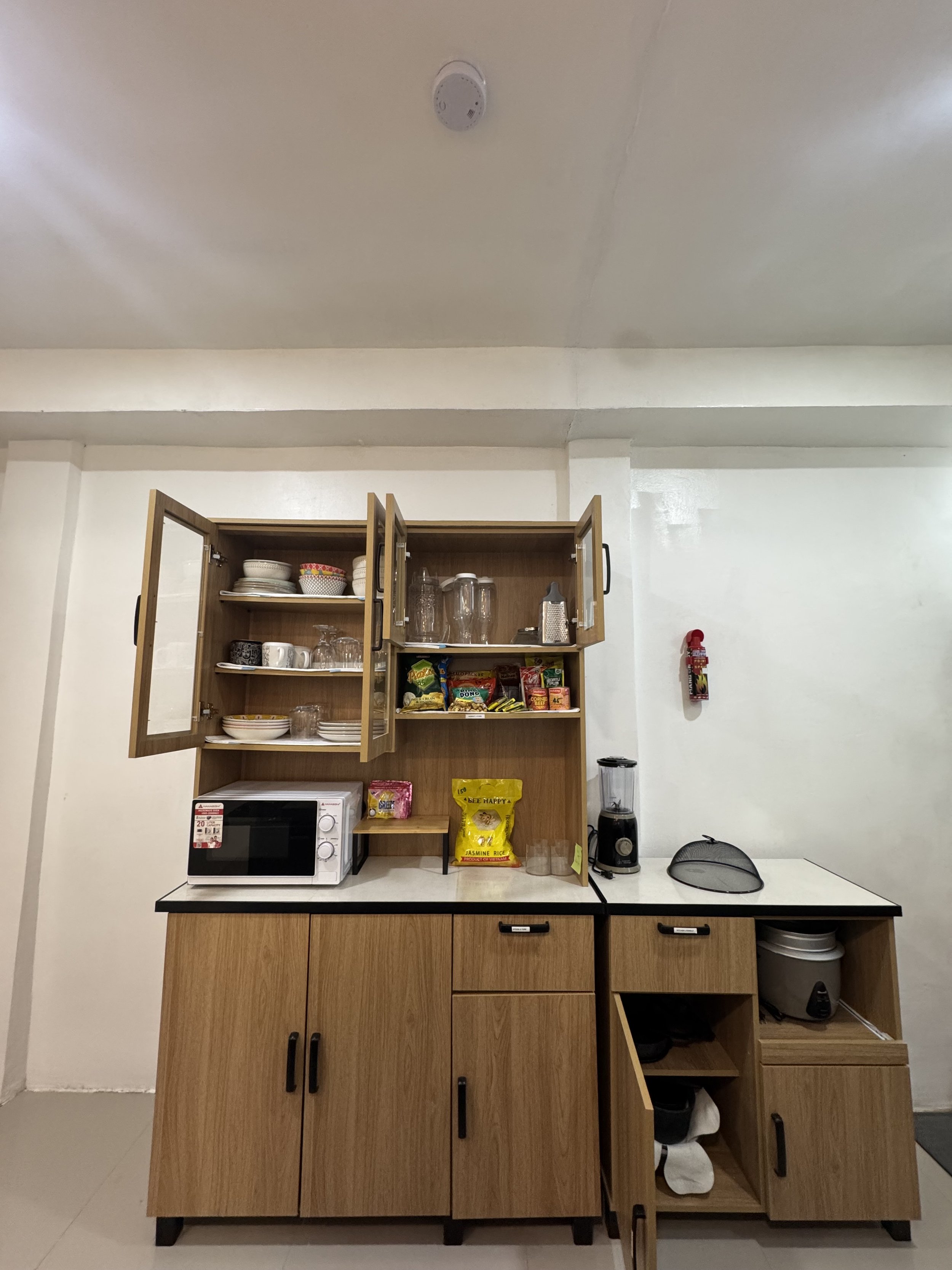 Kitchen cabinet with open doors displaying dishes and glasses, microwave oven, blender, and shelves with snack packets, set against a white wall and ceiling.