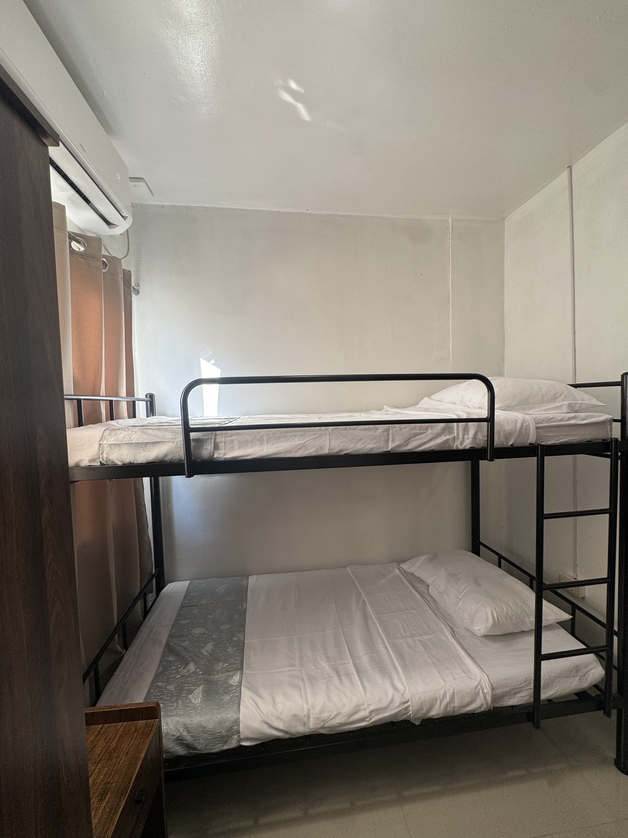 A bedroom with a bunk bed, white bedding on both beds, a window with brown curtains, an air conditioning unit, and a small wooden shelf.