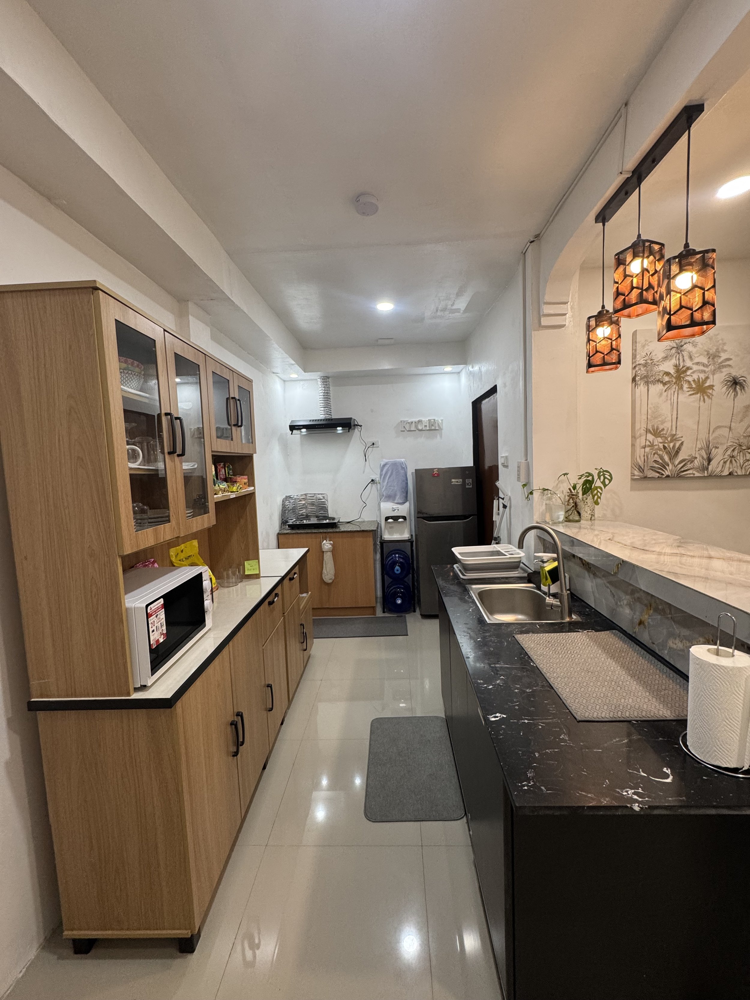A modern kitchen with wooden cabinets, a black marble countertop, and pendant lights with geometric patterns. There is a small microwave oven on the counter, a refrigerator, a sink, and various kitchen appliances and decor.