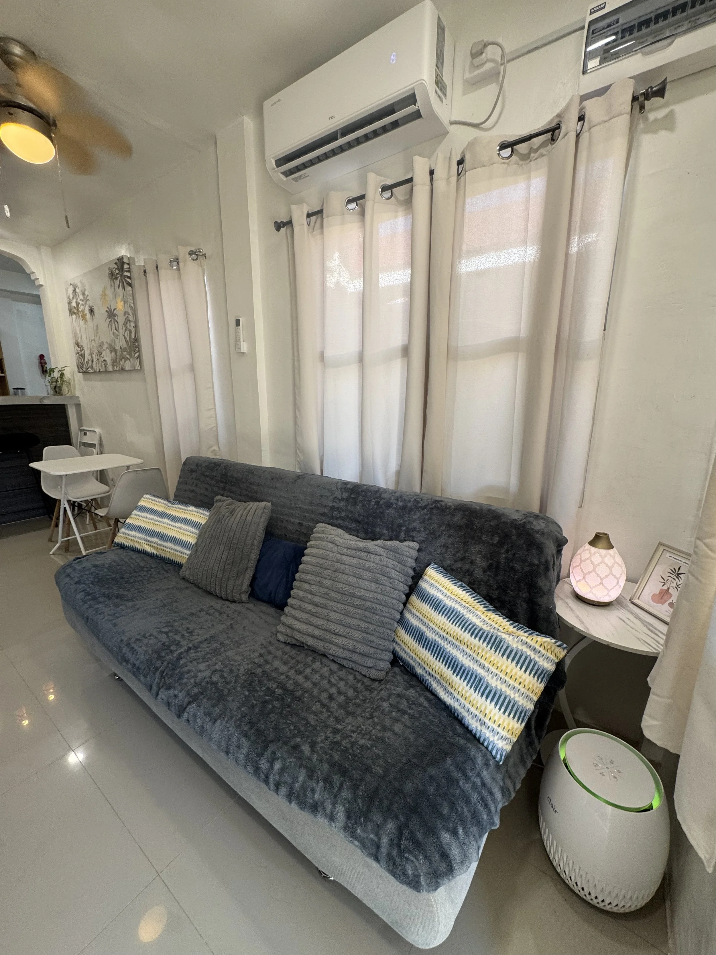 A living room with a grey velvet sofa, decorated with several pillows including striped and grey textured ones. There is a small round white side table next to the sofa, with a decorative lamp and framed picture on it. Behind the sofa, white curtains