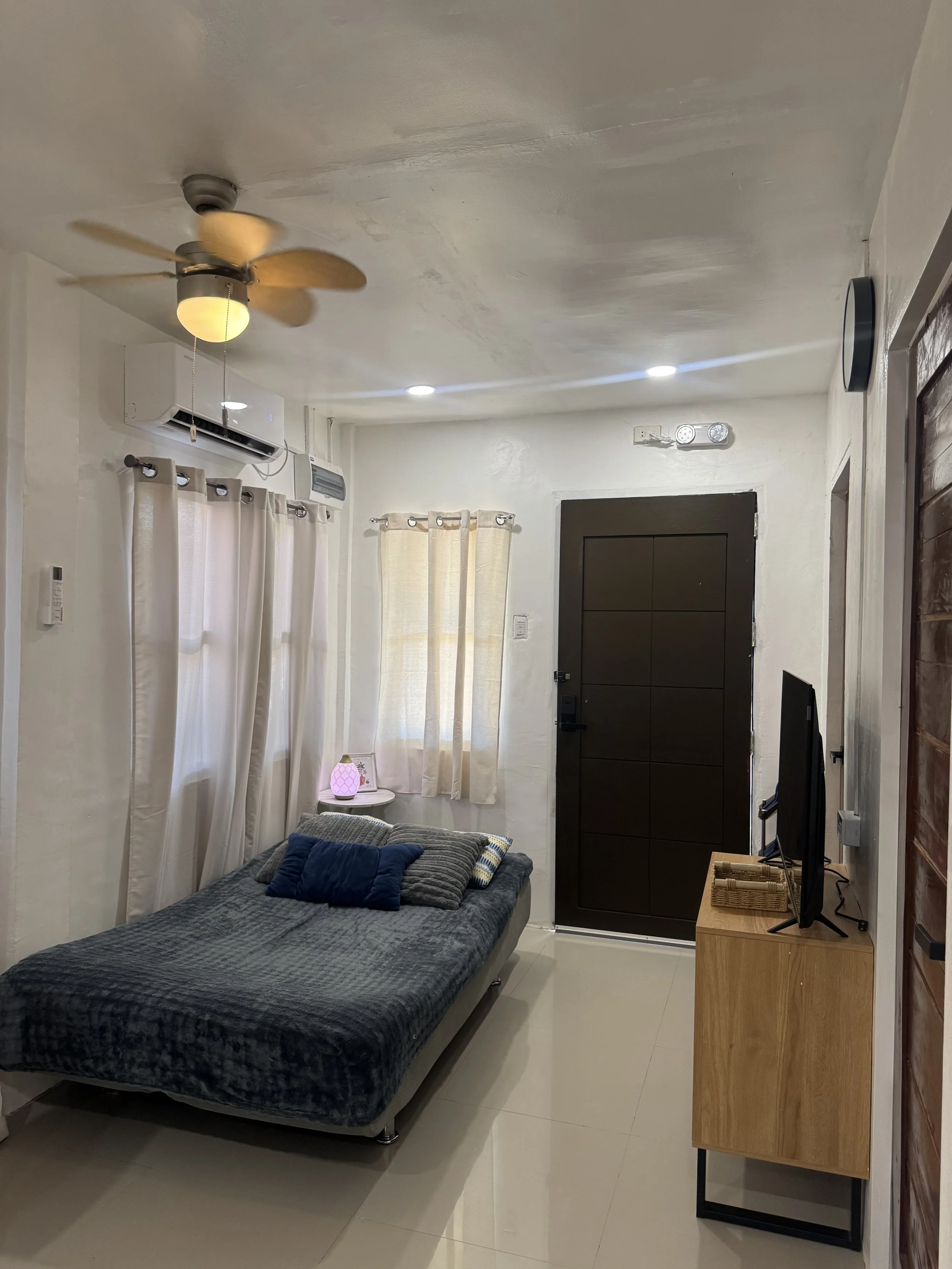 A small, cozy living room with a bed, a television on a wooden stand, an air conditioning unit, curtains on the windows, and a ceiling fan with a light. There is a lampshade on a small table next to the bed.