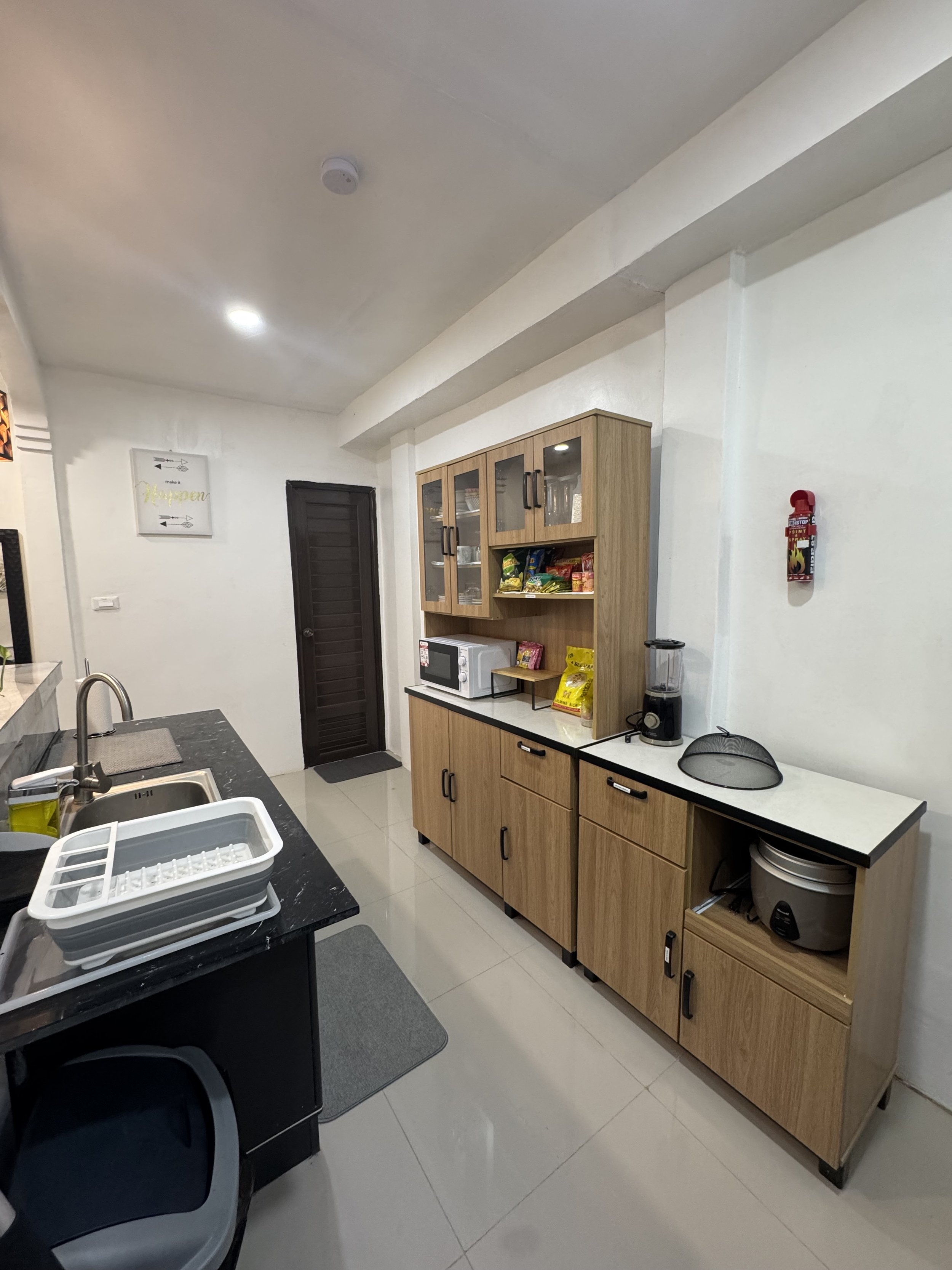 A modern kitchen with wooden cabinets, a microwave, blender, snacks, and a sink. There is a gray rug on the floor and a wall with a sign that says 'happen' with arrows pointing in both directions. A black door is visible in the background.