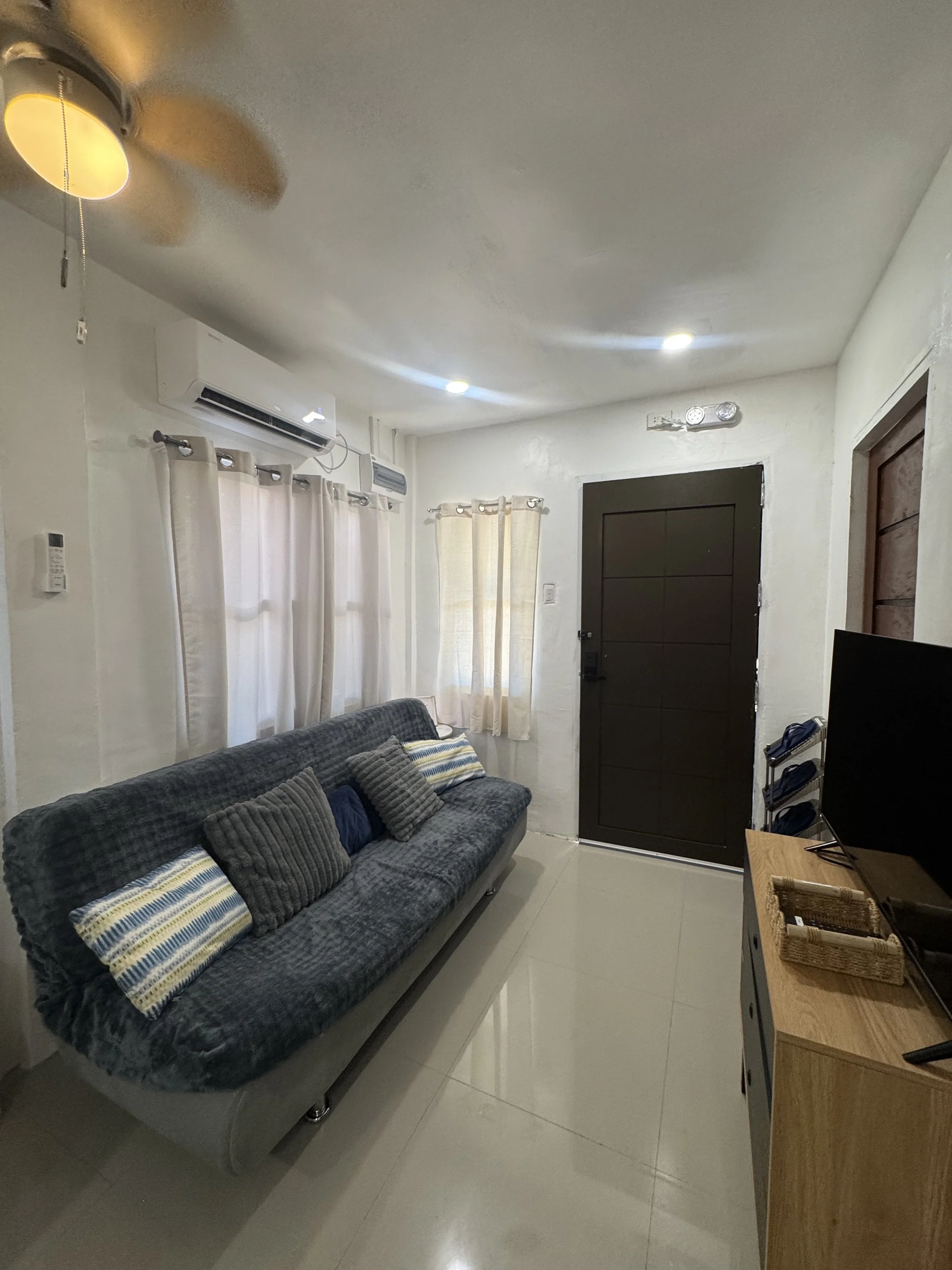 Living room with dark gray sofa with striped pillows, white curtains on windows, black front door, wall-mounted air conditioning unit, ceiling fan with lights, tv on wooden stand, small shelf with baskets, and beige tiled floor.