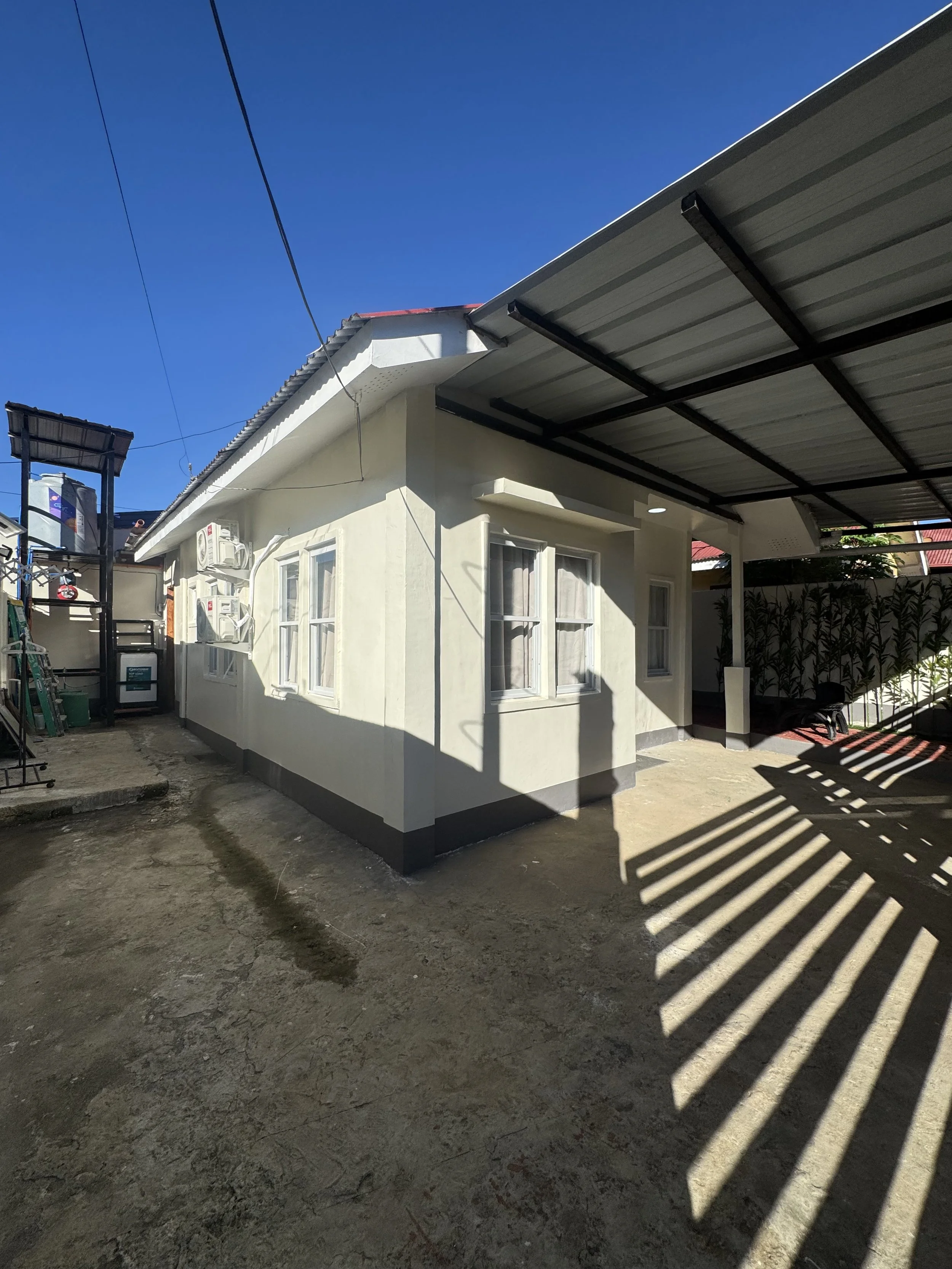 A residential house with a beige exterior, white window frames, and a metal roof. The house has air conditioning units mounted on the wall. There is a covered carport on the right side and outdoor equipment and plants on the left. The ground is concr