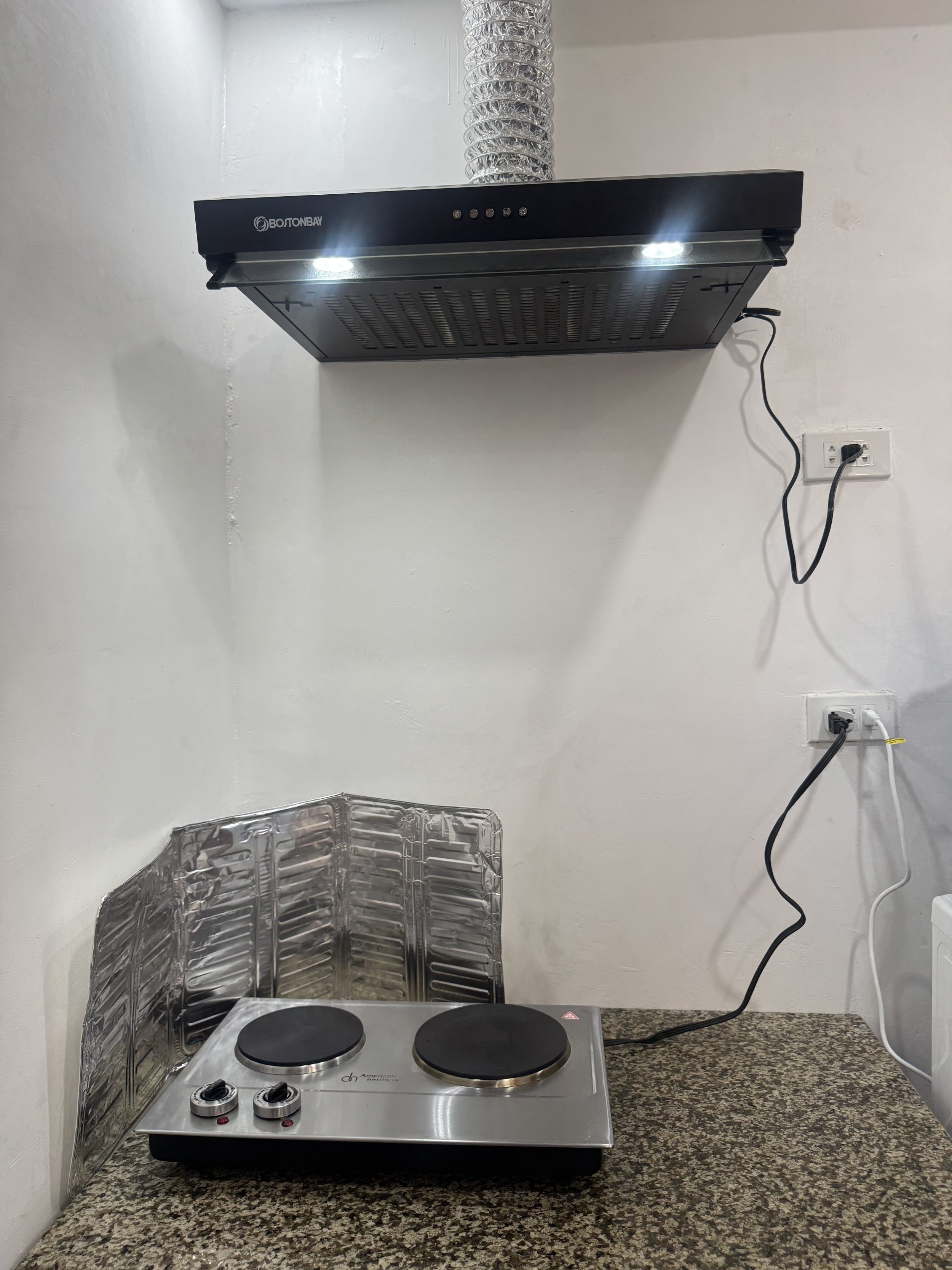 Kitchen countertop with portable electric stove with two burners, a back wall-mounted range hood with lights, and two electrical outlets with cords plugged in.