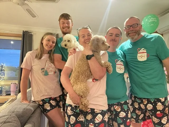 Family celebrating a holiday party with Christmas-themed pajamas and a green balloon, posing together with two dogs inside a living room.