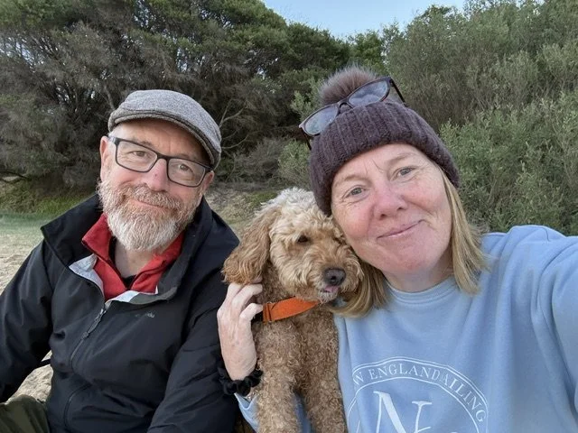 A smiling couple with glasses, dressed warmly, sitting outdoors with a fluffy brown dog between them. The woman is wearing a gray beanie and sunglasses on her head, and the man has a cap and beard. Trees and bushes are in the background.