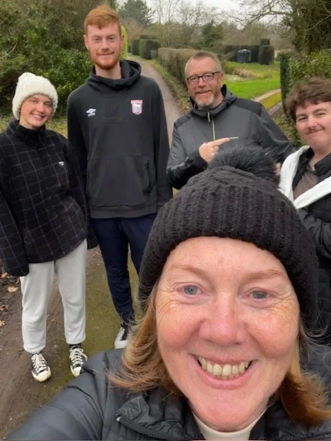 A group of five people taking a selfie outdoors, smiling. One person in the foreground, a woman, is holding the camera. The others are standing behind her on a grassy path, with trees and bushes in the background.
