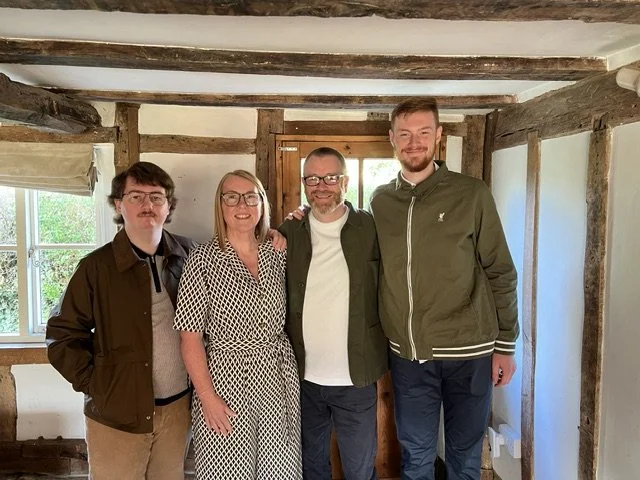 Four people standing together indoors, smiling. The group includes two men with glasses, a woman in a polka dot dress, and a taller man in a green jacket. They are in a rustic room with exposed wooden beams and a small window behind them.