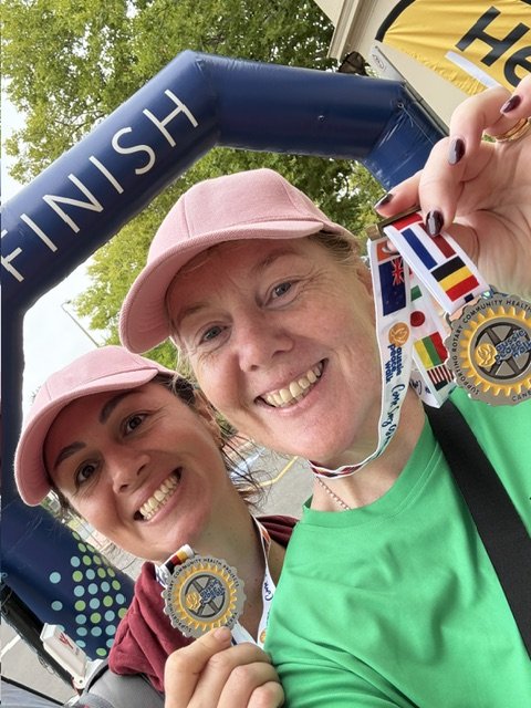 Two women in pink caps smiling and holding medals at a finish line arch after a race.