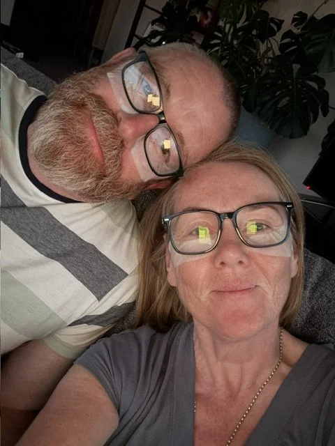 A man and woman wearing glasses lying close together, taking a selfie indoors with plants and a computer in the background.