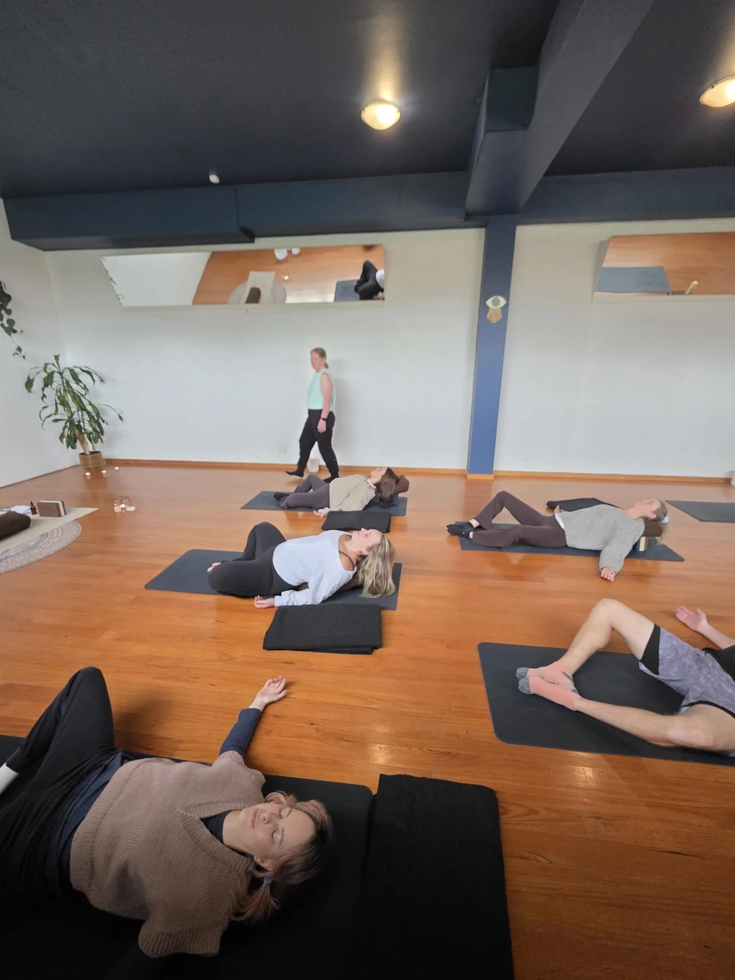 People practicing yoga in a studio, lying on mats with eyes closed, some resting on pillows, in a calm and peaceful environment.