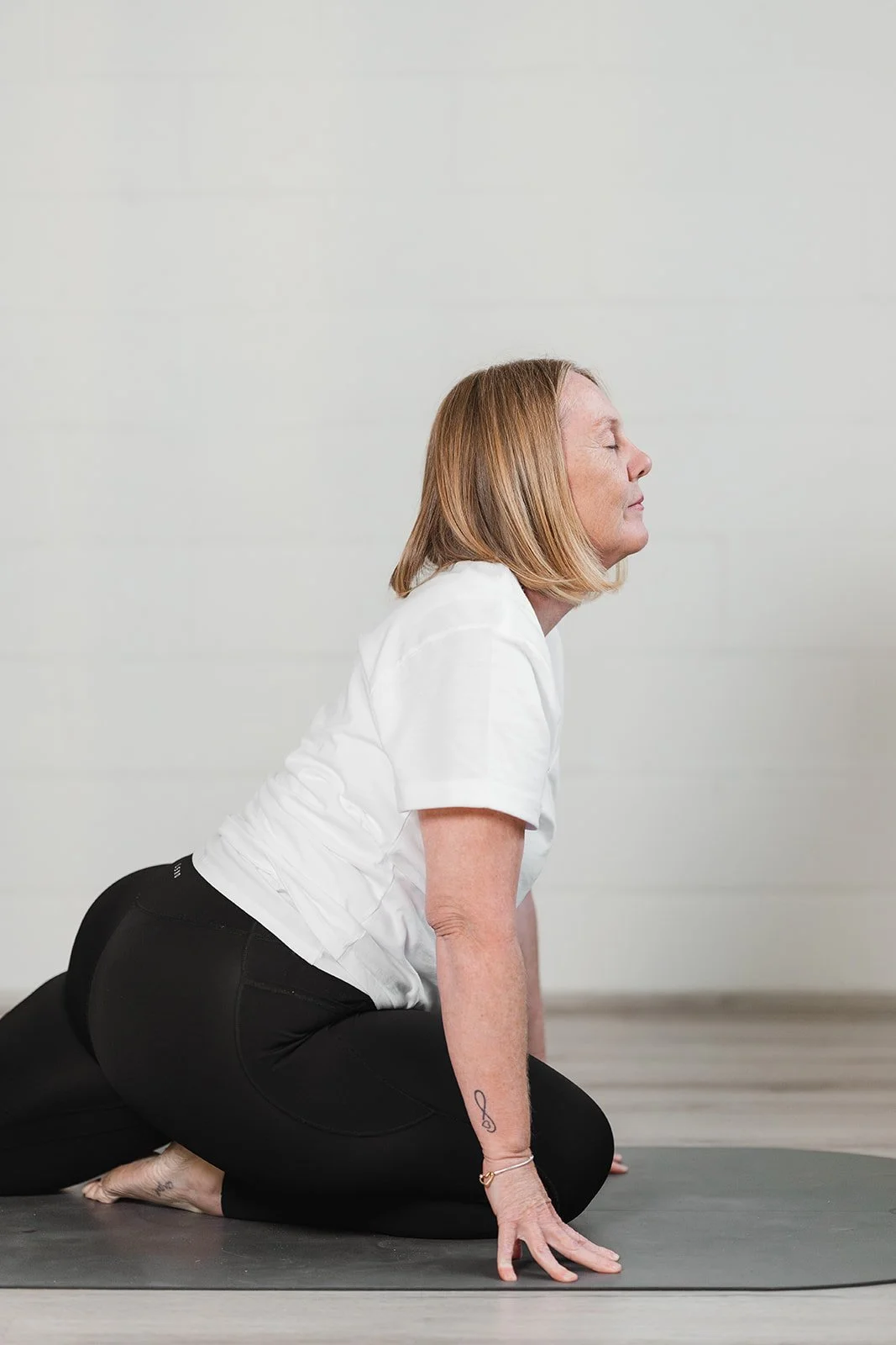 A woman with blonde hair practicing yoga on a black mat, in a low lunge pose, wearing a white shirt and black pants.