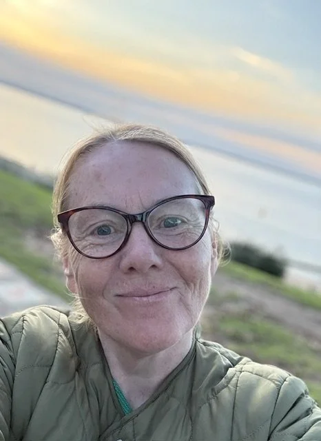 A woman with red hair, glasses, and freckles taking a selfie outdoors during sunset, with a grassy field and a cloudy sky in the background.