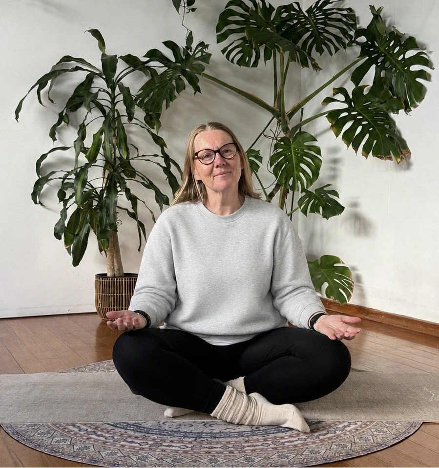 A woman sitting cross-legged on a yoga mat indoors, gesturing with her hands in a meditative pose, with large tropical plants behind her.