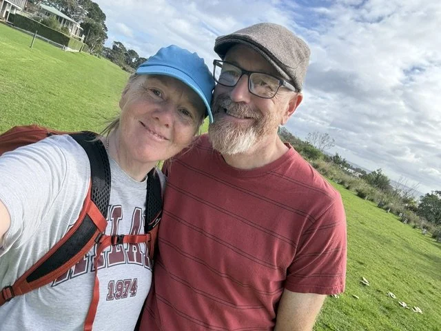 A smiling woman wearing a blue cap and gray t-shirt with red lettering, standing next to a smiling man with glasses, a gray cap, and a red striped t-shirt, outdoors on a grassy field with trees and cloudy sky in the background.
