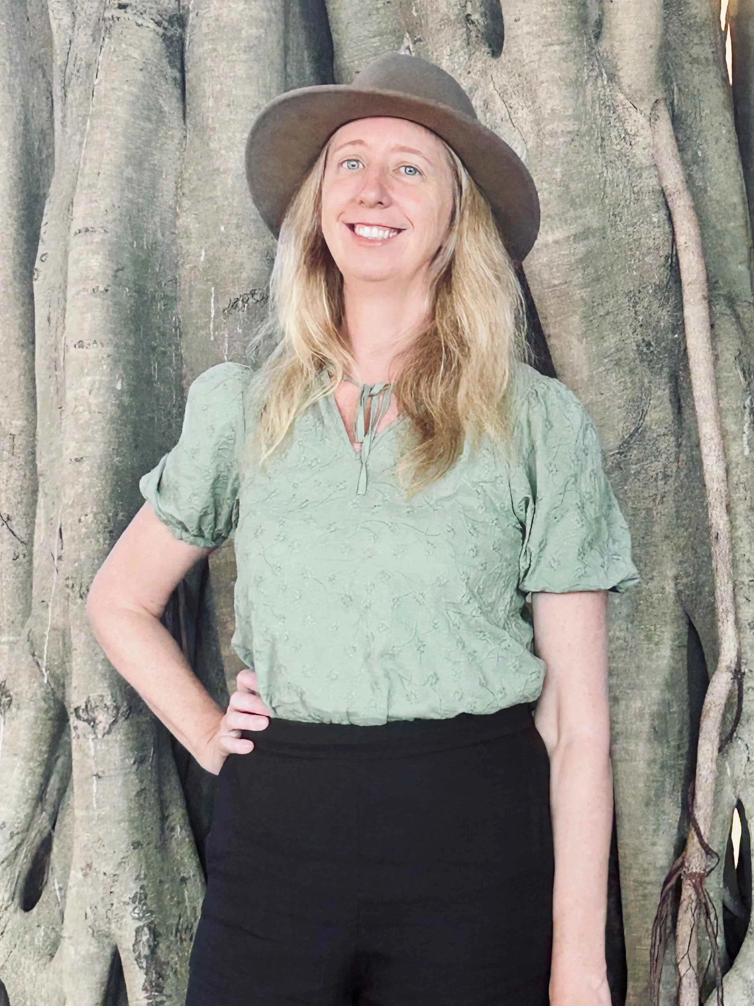 Dr Debbie Gleeson wearing a wide-brimmed hat, green blouse, and black skirt, standing outdoors near a large tree with twisted roots.