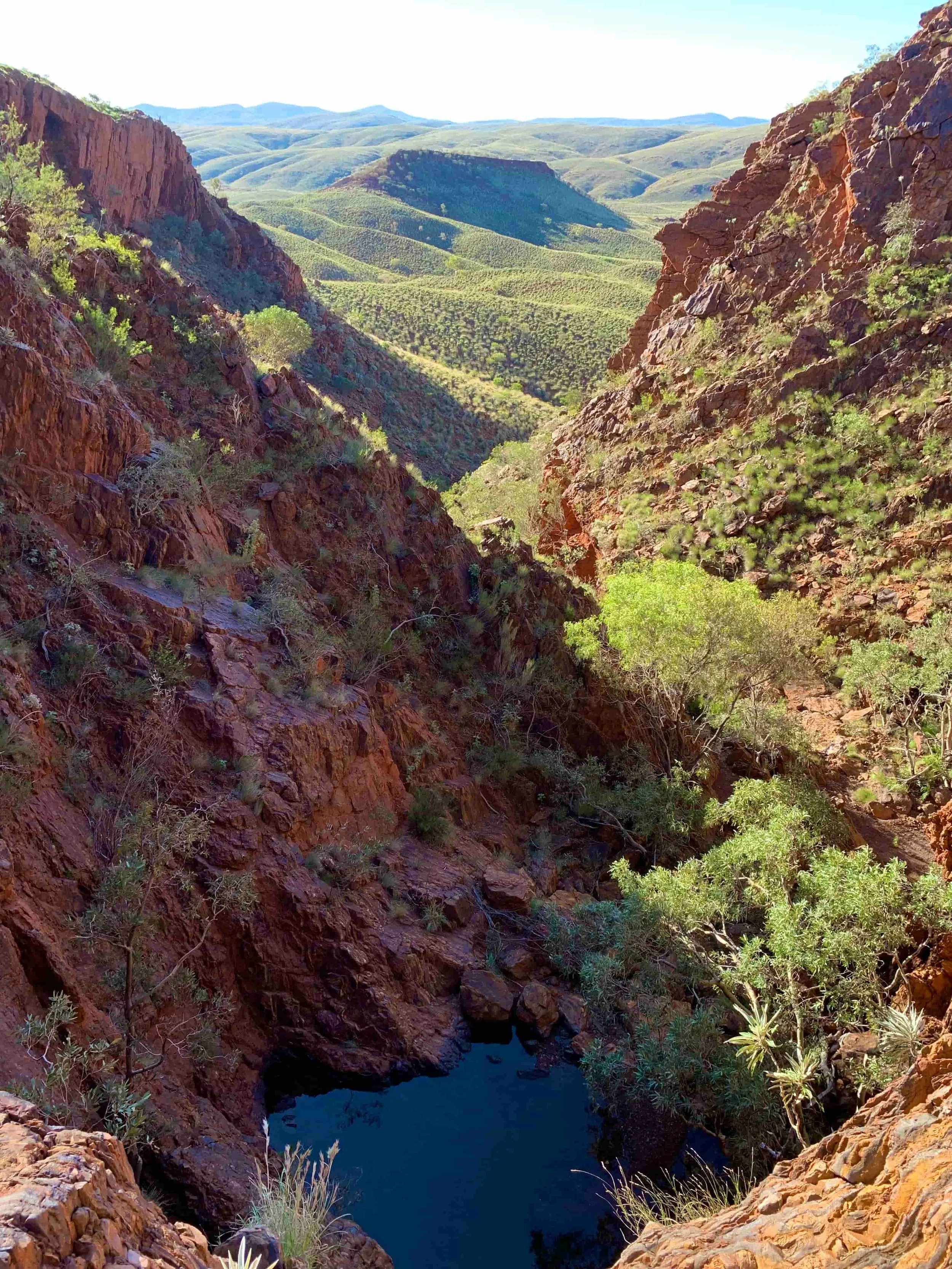 A deep canyon with red rocky cliffs on either side, a narrow water stream at the bottom, and green hills and mountains in the background under a clear blue sky.