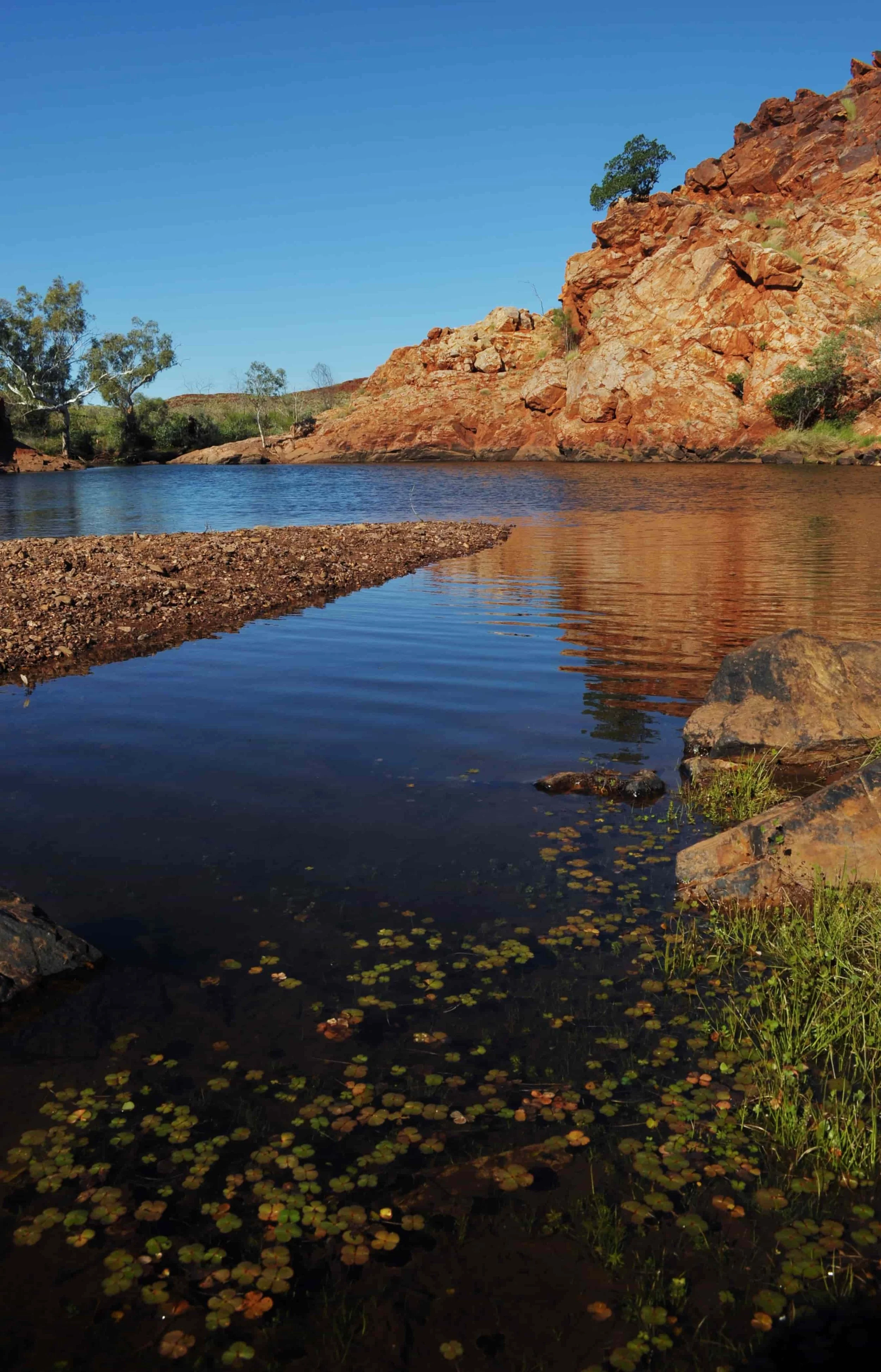 A river with calm water reflecting the blue sky, surrounded by reddish-brown rocky cliffs and sparse green vegetation.
