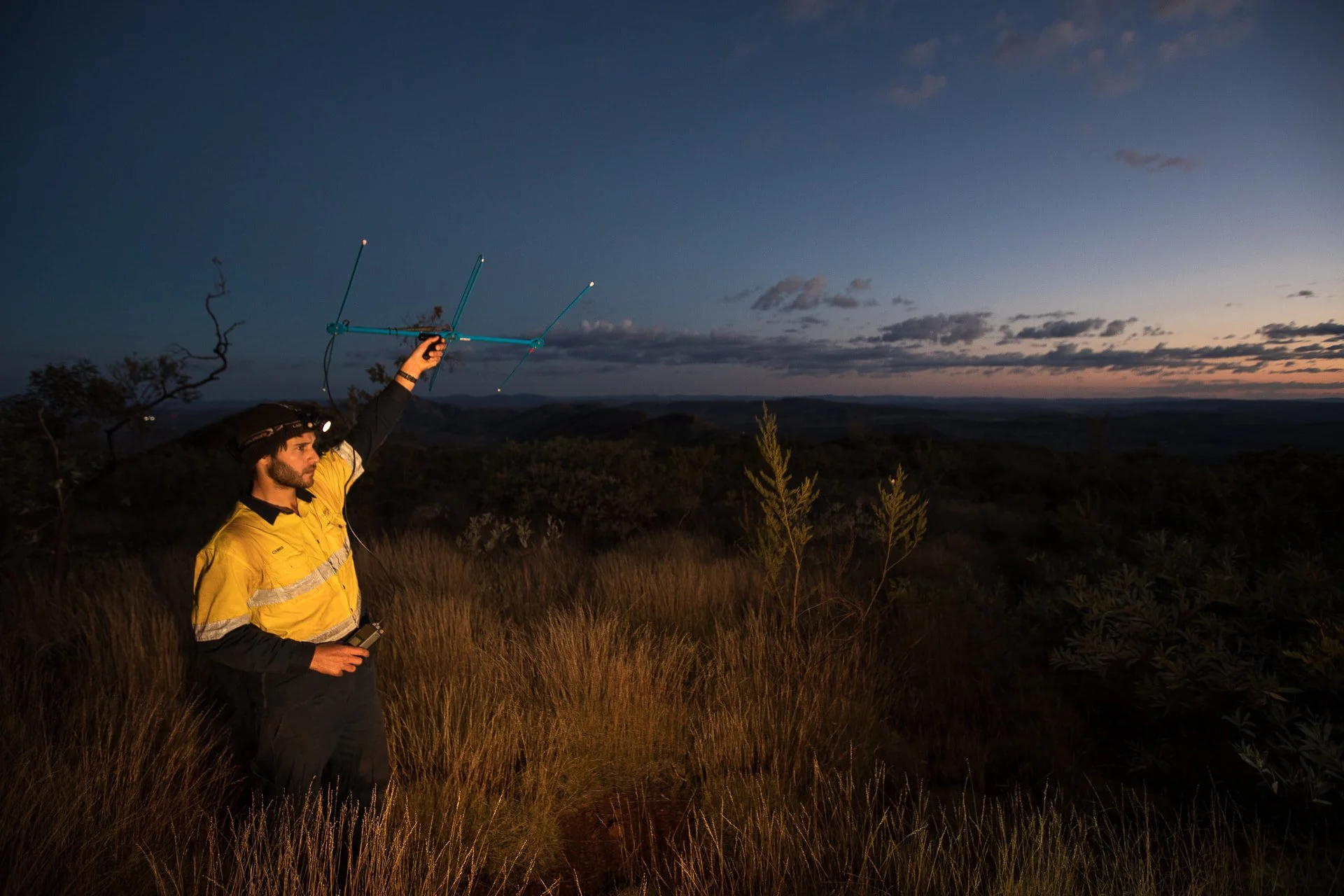 Environmental Surveyor Chris Knuckey wearing a yellow jacket with reflective stripes holds a directional antenna in an open field during dusk, with a landscape and a partly cloudy sky in the background.