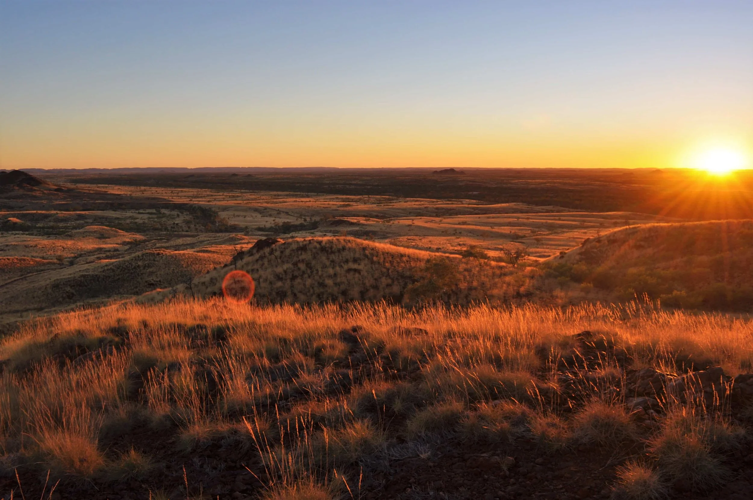 Sunset over a vast, open landscape with rolling hills and dry grass in the foreground.
