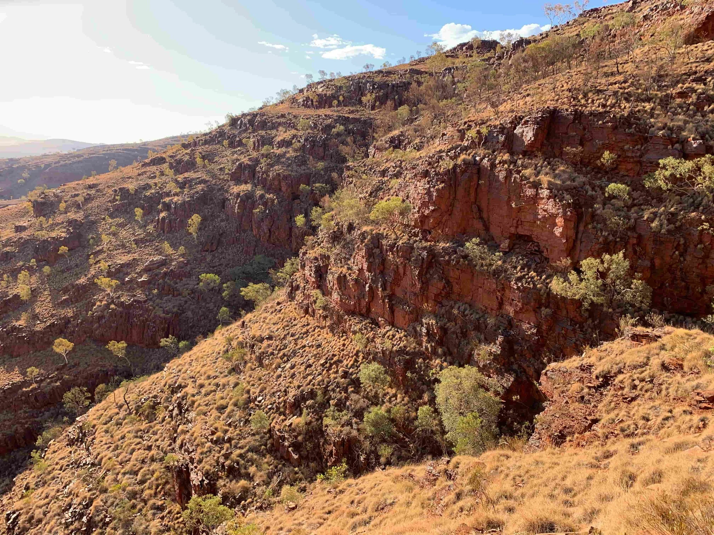 A rugged Australian hillside terrain with reddish-brown rocks and sparse vegetation under a partly cloudy blue sky.