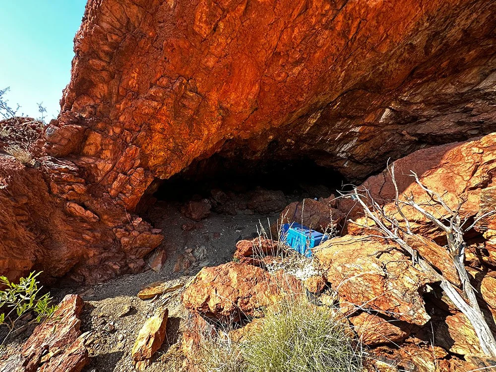 A rocky red-orange cave opening with a blue cooler inside and sparse desert vegetation outside.