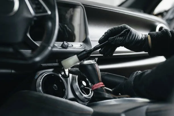 Person cleaning the interior dashboard of a car with a brush, wearing black gloves.