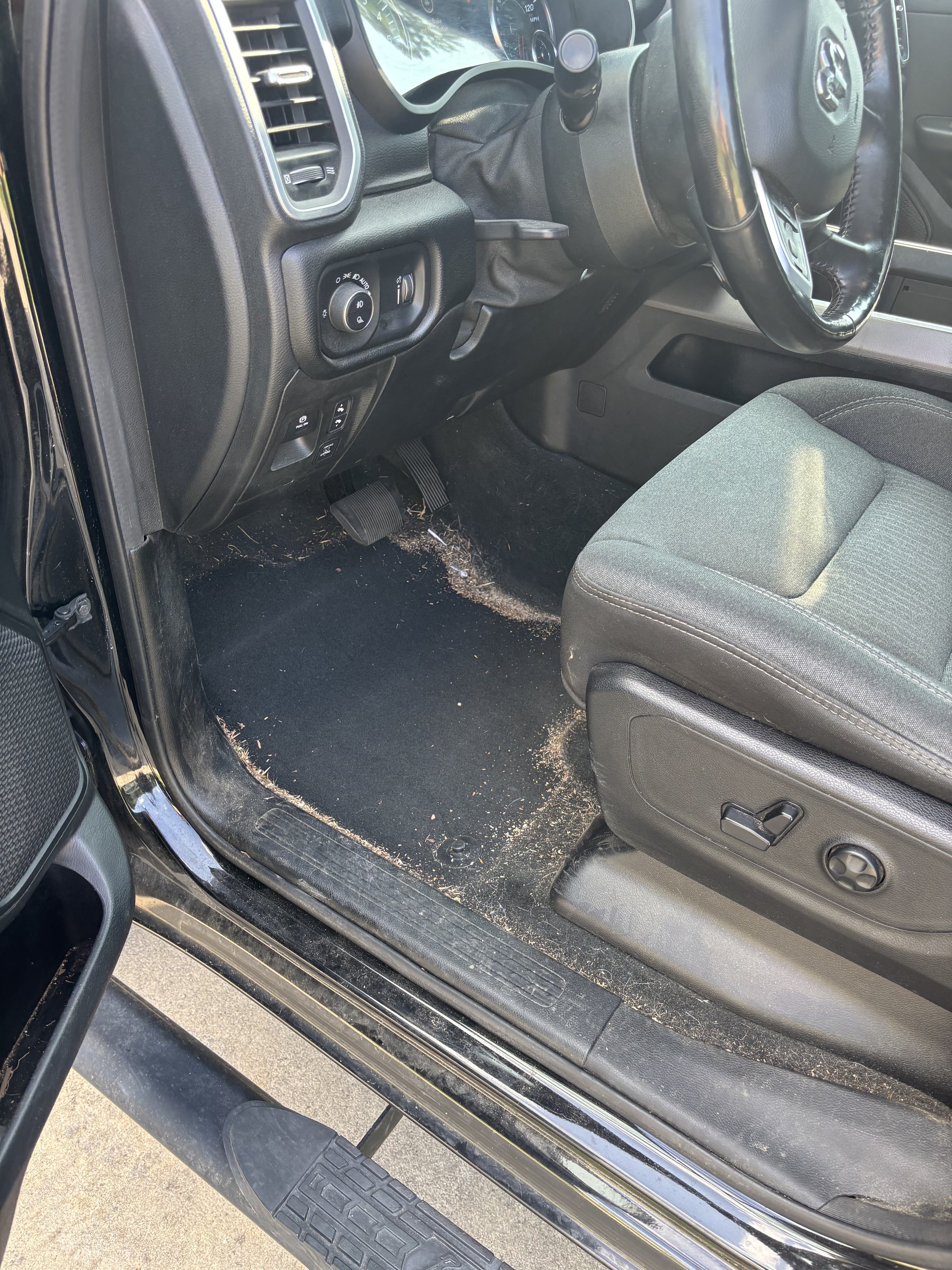 Inside a vehicle with a black interior, showing the driver's seat, steering wheel, dashboard, and floor area with some dirt and debris.