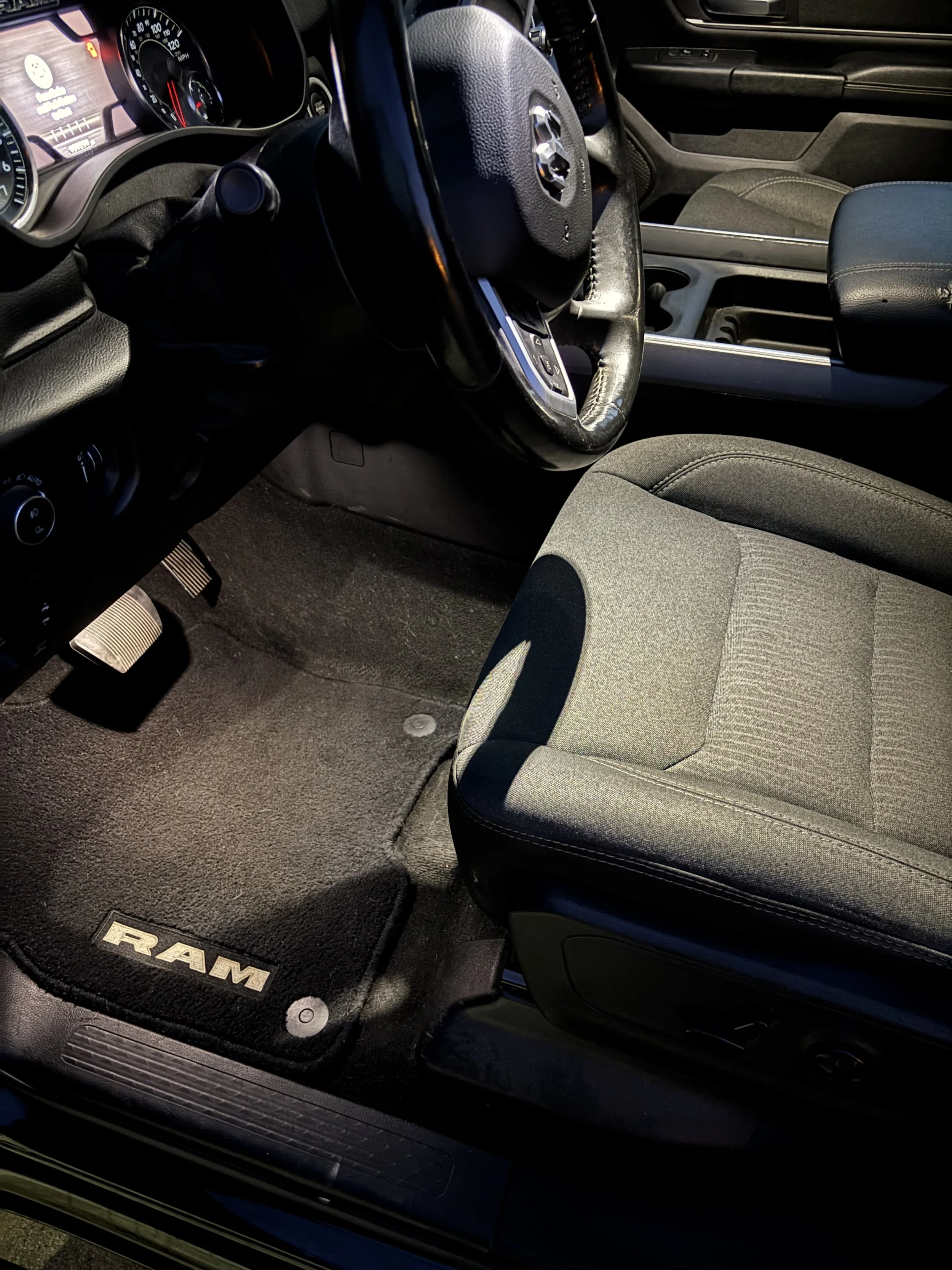 Interior of a pickup truck showing the driver's seat, steering wheel, dashboard with speedometer and touchscreen display, pedals, and a floor mat with the RAM logo. The seat is gray fabric, and the dashboard is black.