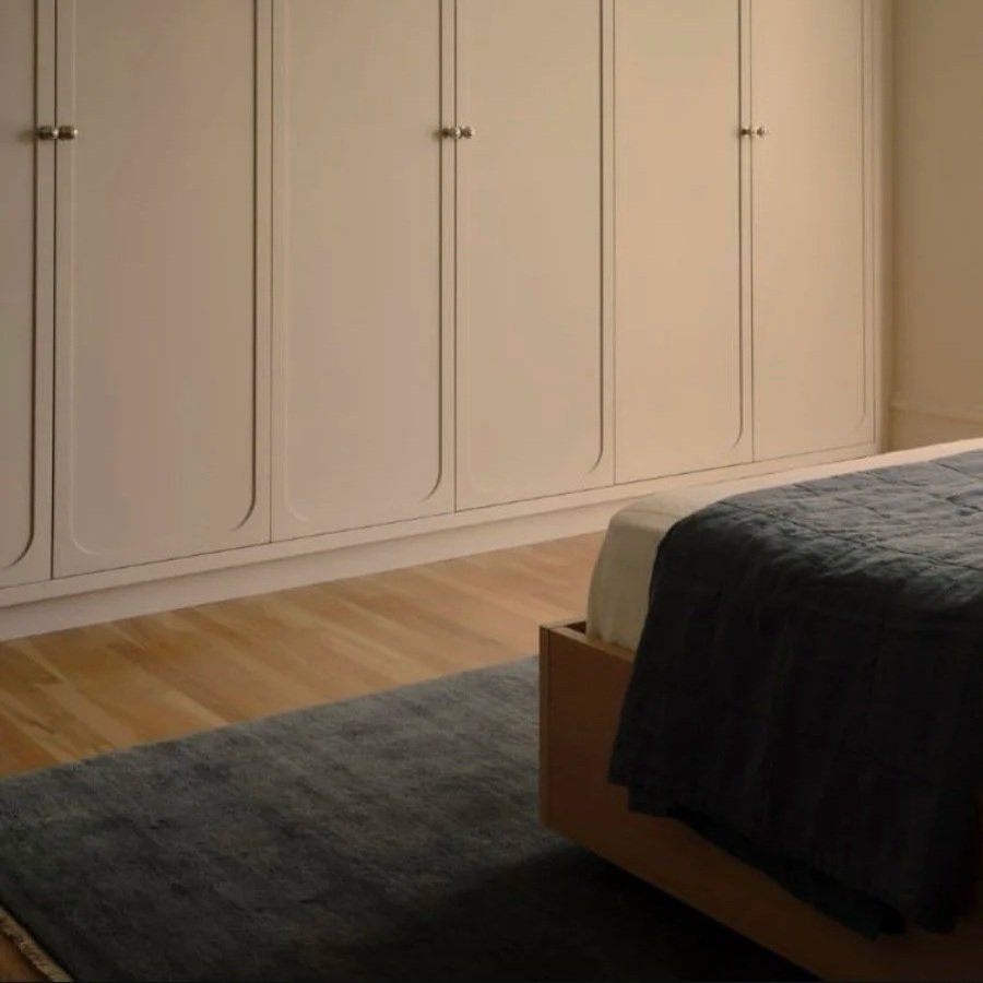 A bedroom with a large white closet, part of a bed with dark bedding, and a gray rug on light wooden flooring.
