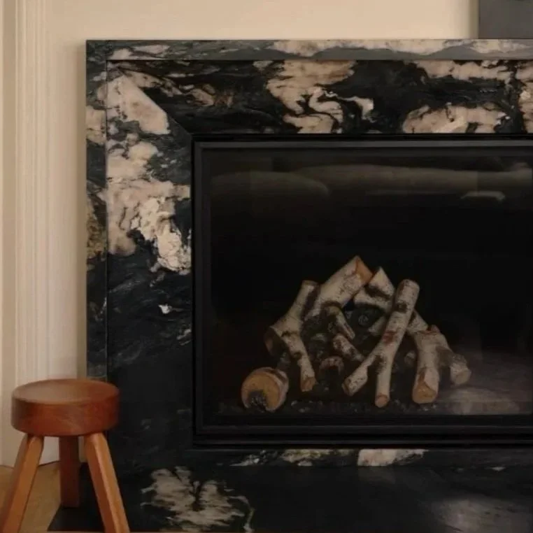 A fireplace with a black and white marble mantel, and logs inside. To the left, a small round wooden stool with three legs.