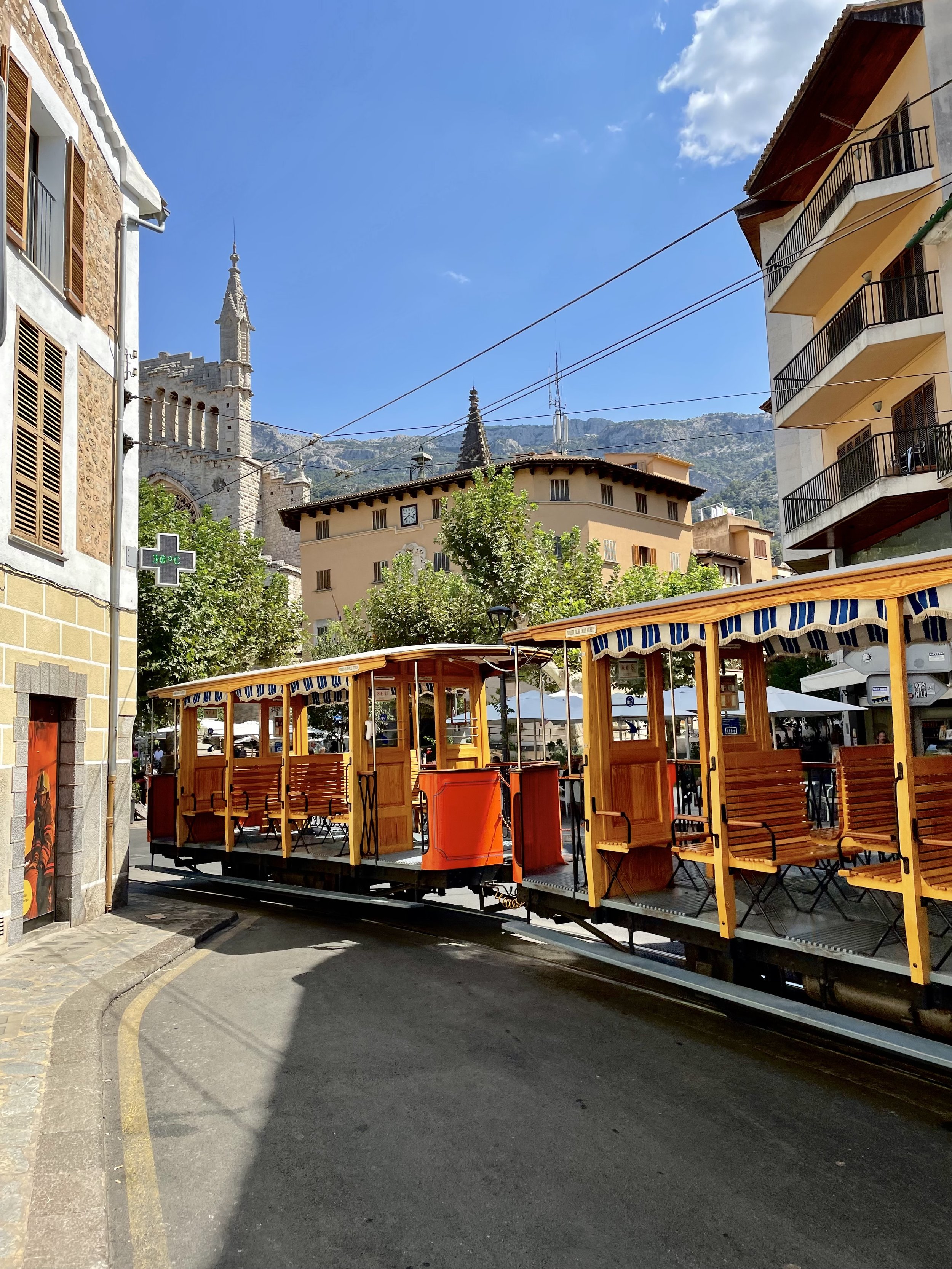 A street scene with a small train ride with orange and yellow carriages, buildings with balconies, a church tower, and mountains under a partly cloudy blue sky.