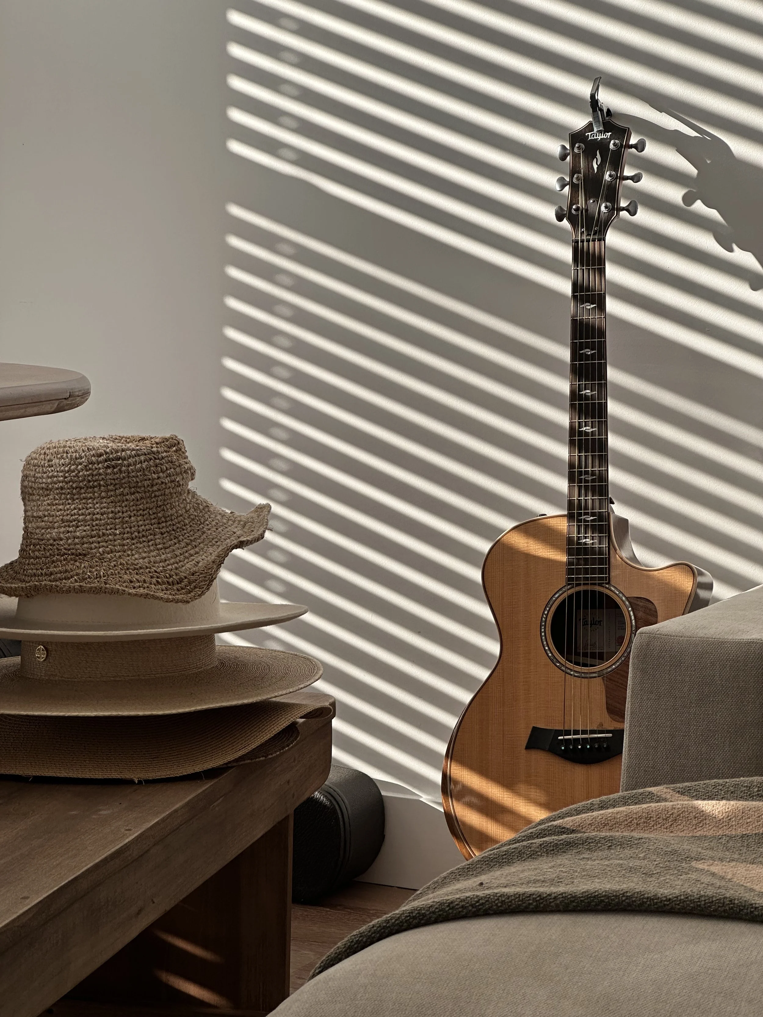 An acoustic guitar leaning against a wall with shadow stripes from a sun-lit window, a woven hat on a dark natural wood bench, and part of a beige sofa in a cozy room.