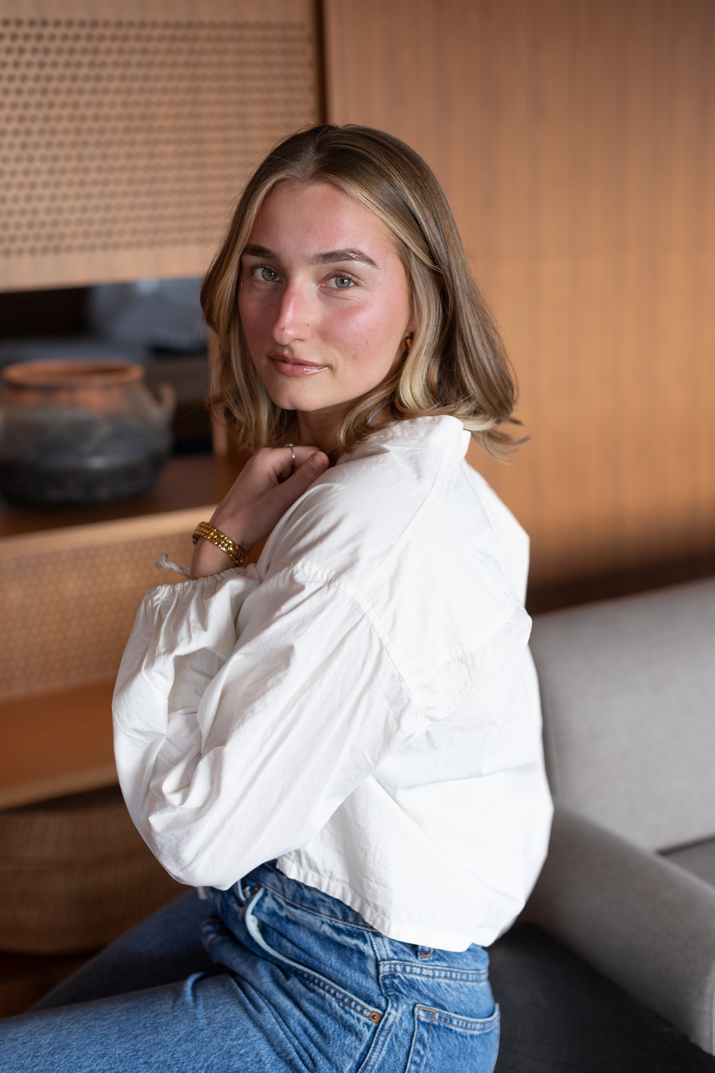 A young woman sitting on a sofa, wearing a white shirt and blue jeans, with a brown wooden panel and a decorative ceramic pot in the background.