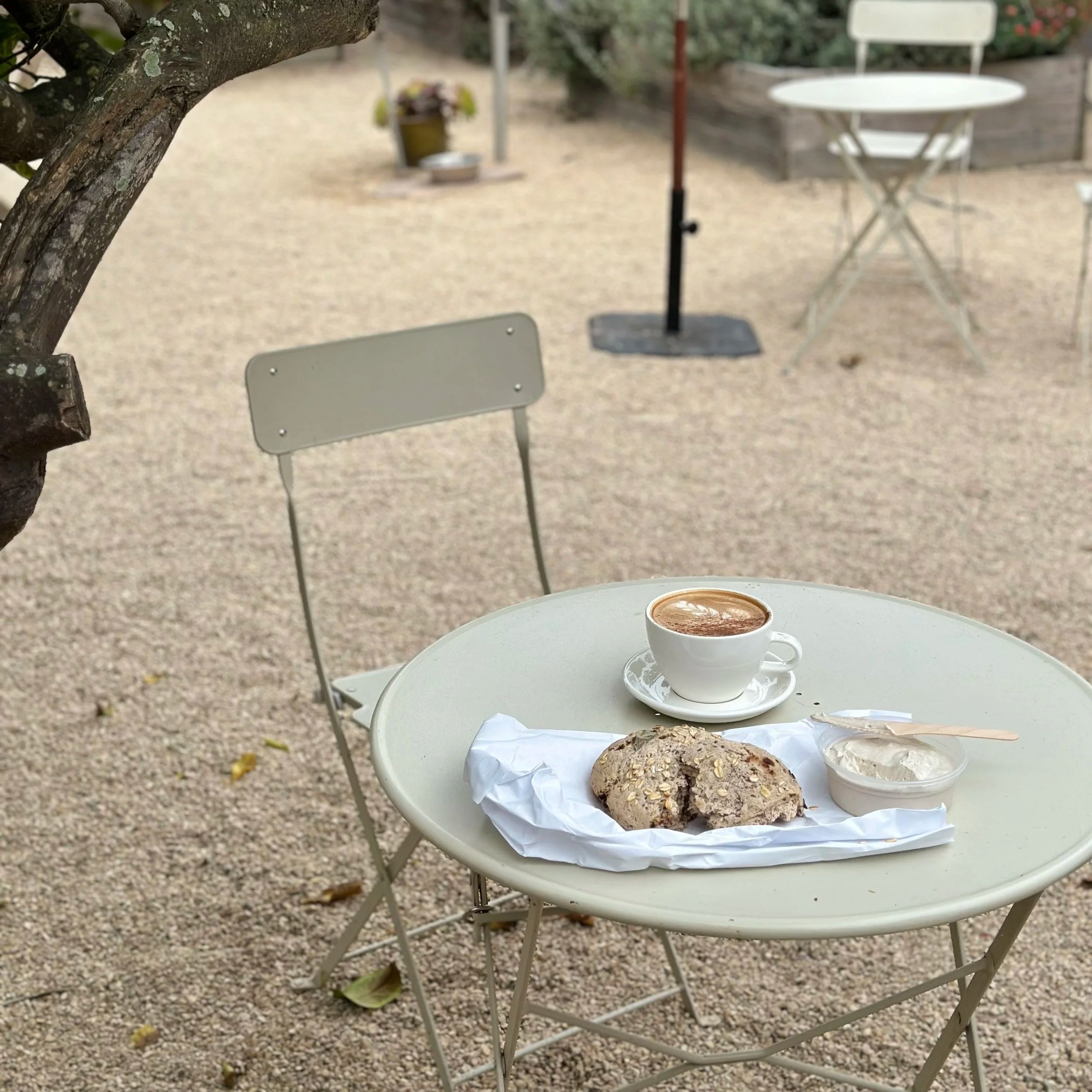 A white table with a cup of hot chocolate, a loaf of bread, and wrapped pastry, set outdoors in a garden or patio with chairs, a tree, and gravel ground.
