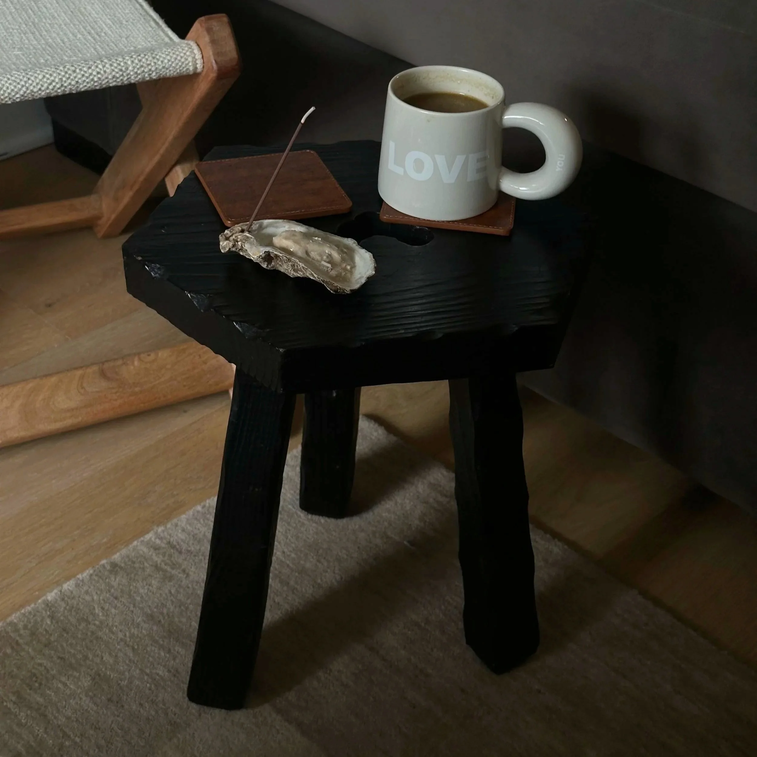 A white ceramic mug filled with coffee on a dark vintage stool next to a brown leather coaster, a custom oyster incense holder and incense burning within in, with a wooden chair and beige wool carpet in the background.