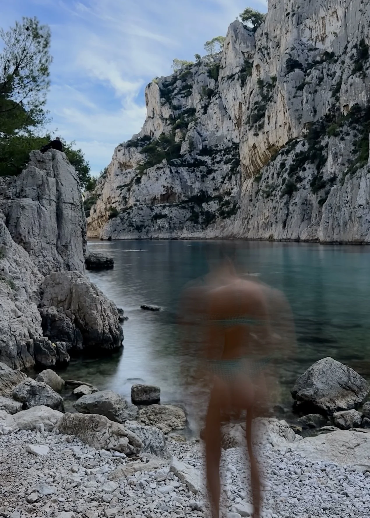 Person walking along rocky shoreline next to calm river or lake, surrounded by towering white rocky cliffs with sparse vegetation, under a partly cloudy sky.
