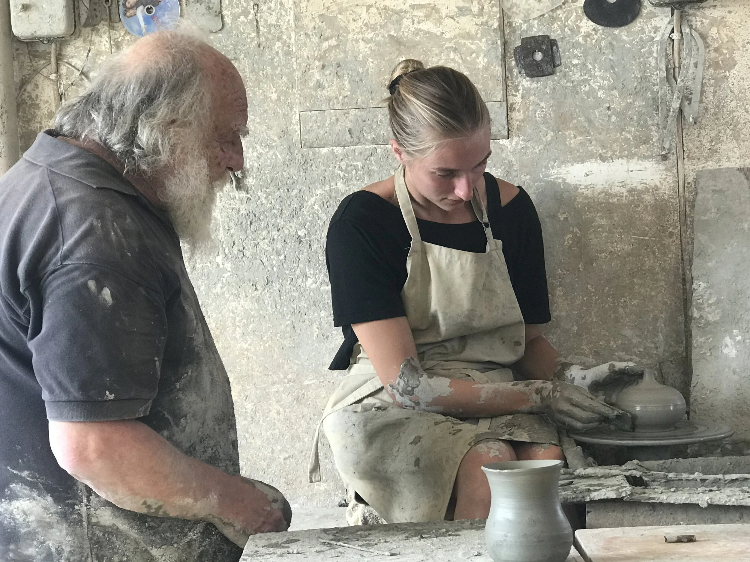 A man with a gray beard and a woman with blonde hair in a bun are working together on a pottery project in a studio with rough, beige walls. The woman is shaping clay on a pottery wheel while the man observes.