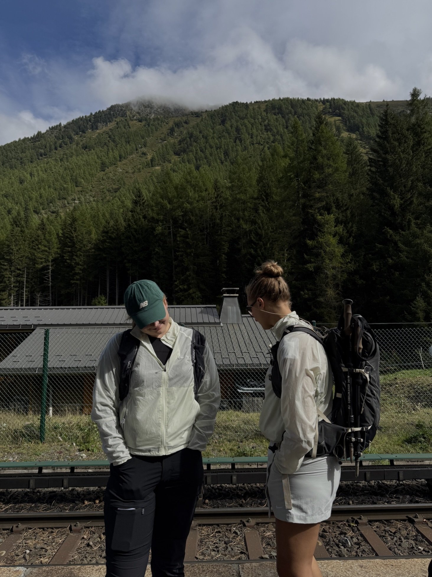 Two women standing on a railroad platform with mountain scenery and a partly cloudy sky behind them. They are wearing outdoor gear and backpacks, looking down at something out of view.