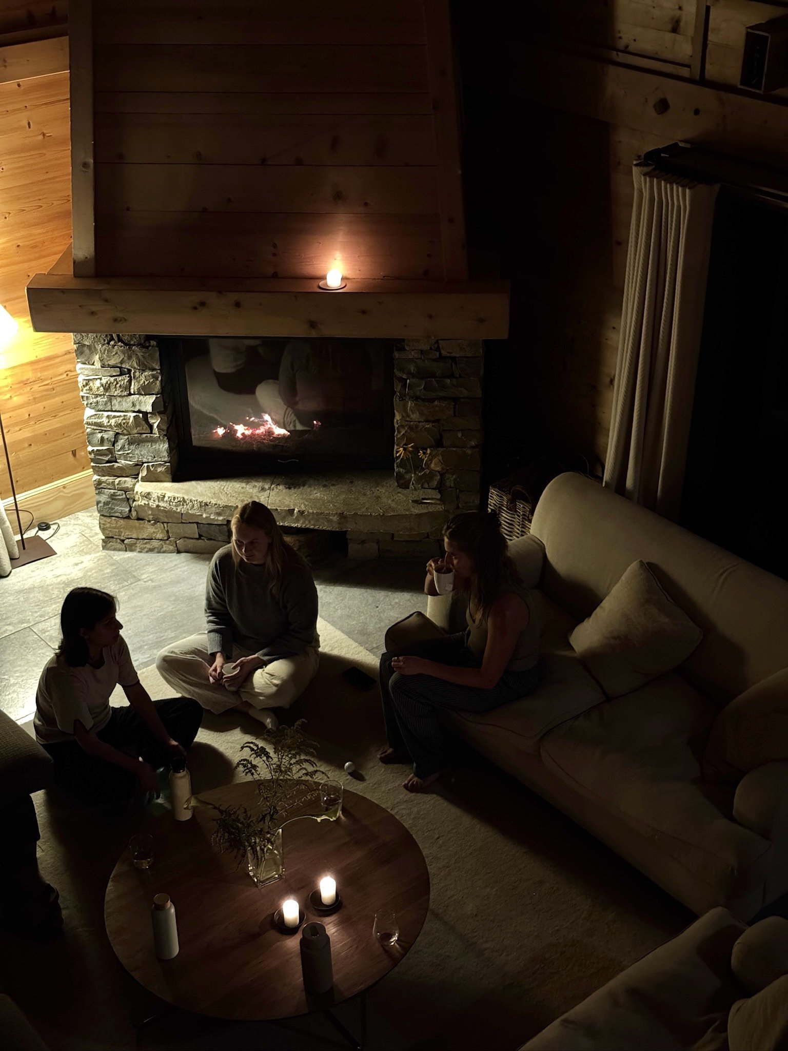 A cozy living room scene in Chamonix, France, with three women gathered on the floor and a sofa, lit by candles with a fireplace and wood-paneled walls in the background.