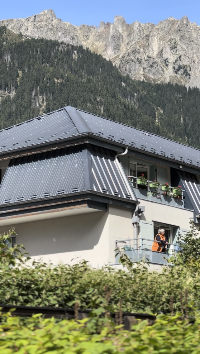 A house with a metal roof and balcony, set against a mountain backdrop with evergreen trees.