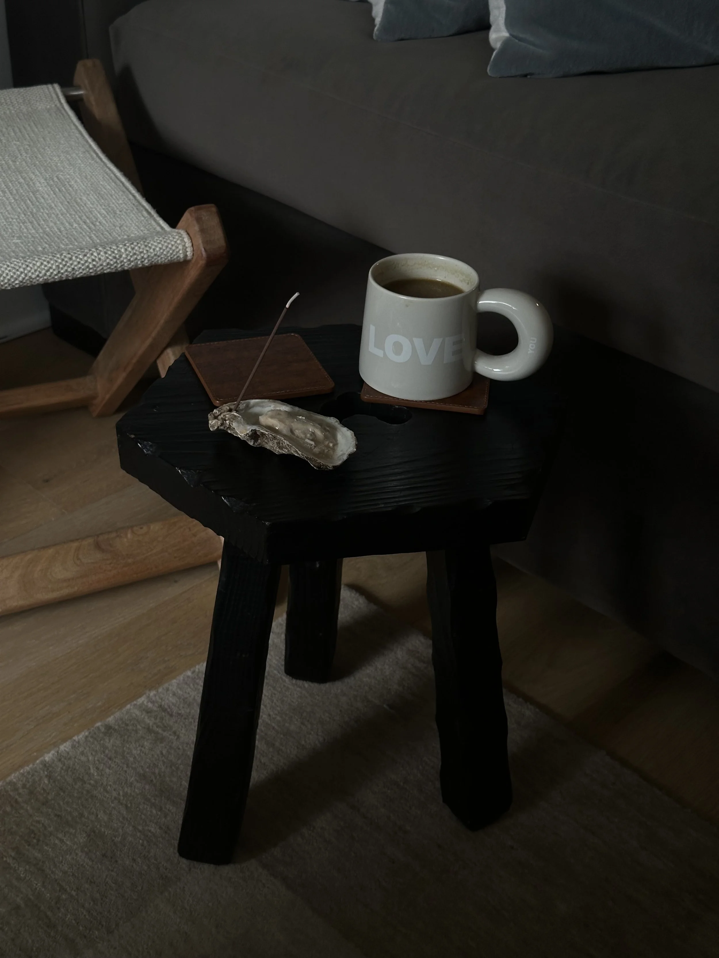 A white ceramic mug filled with coffee on a dark vintage stool next to a brown leather coaster, a custom oyster incense holder and incense burning within in, with a wooden chair and beige wool carpet in the background.