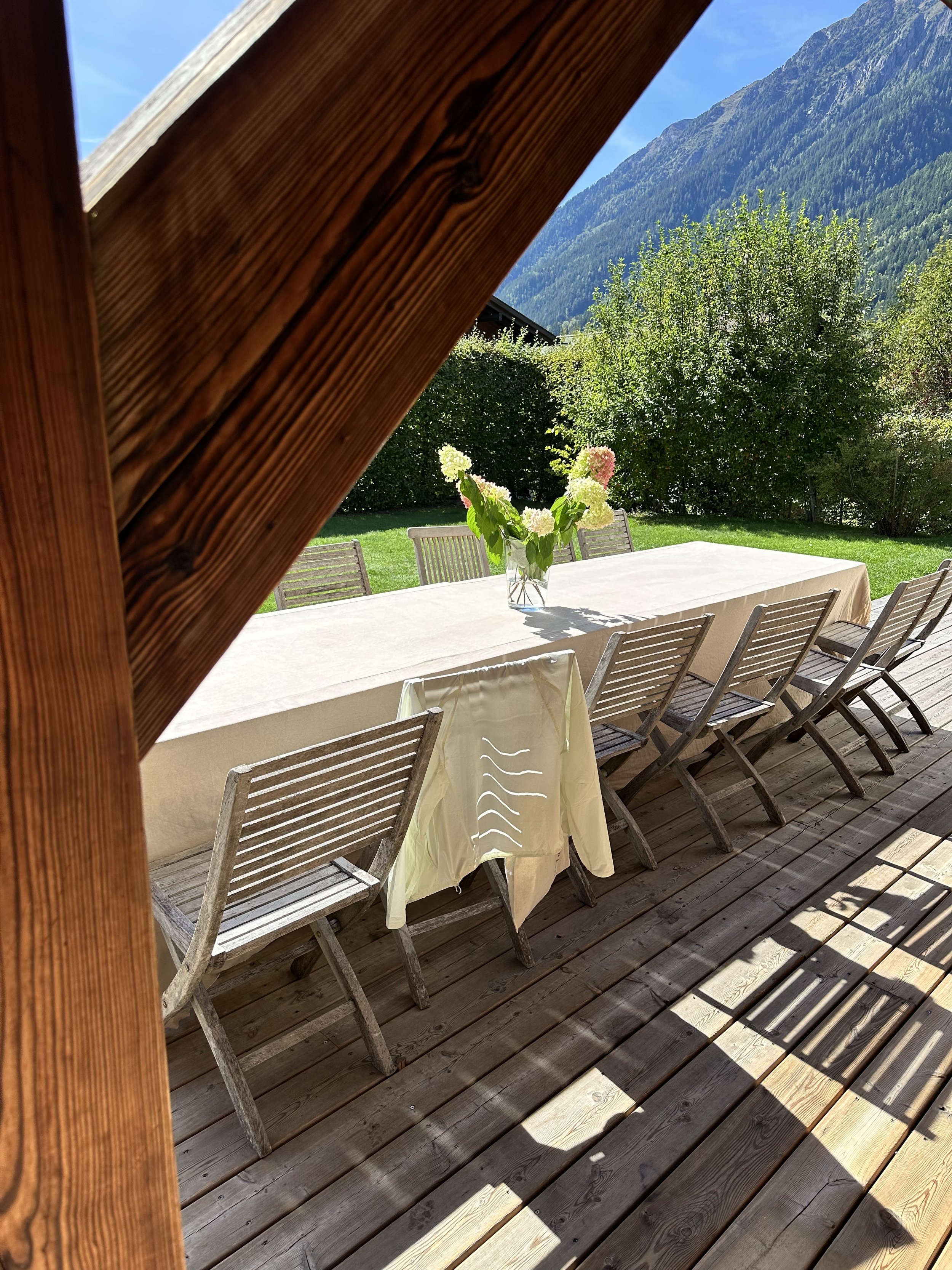 Outdoor dining area with a long table covered with a white tablecloth and surrounded by wooden chairs, a glass vase with white and pink flowers, lush green yard with trees and bushes, mountain backdrop, sunny clear sky, and wooden porch floor.