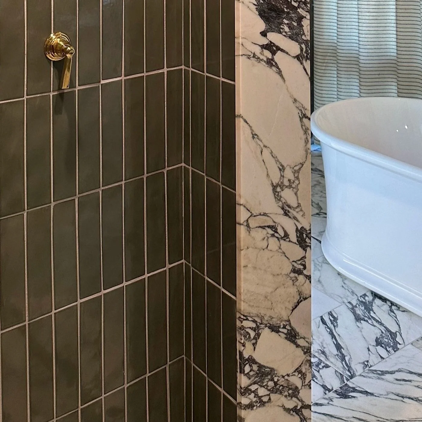 A bathroom corner with dark green tiled shower walls, a marble tiled ledge, and a white bathtub with marble floor and wall tiles.
