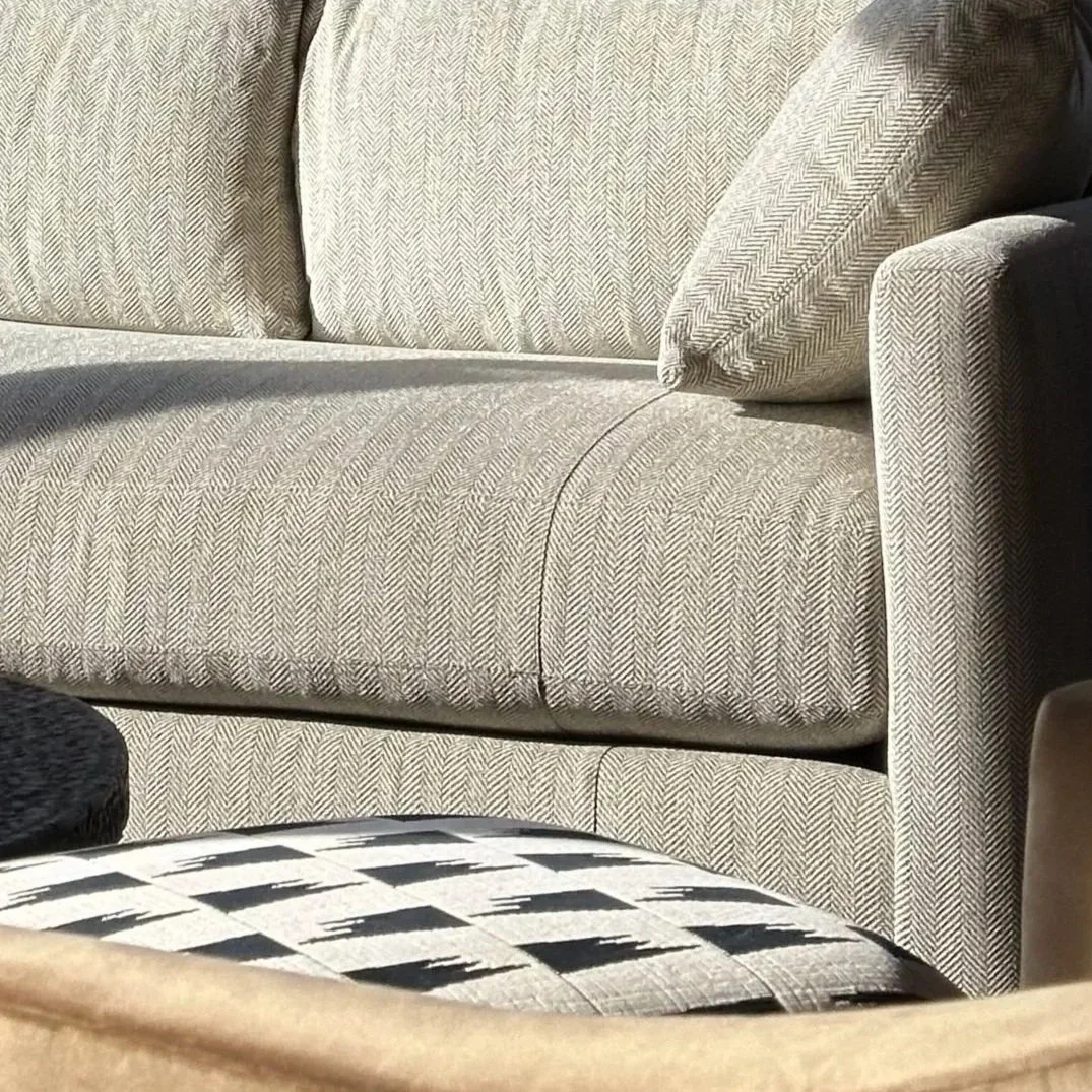 Close-up of a beige upholstered sofa with a herringbone pattern, showing cushions and armrest, with part of a patterned black and white ottoman in front.